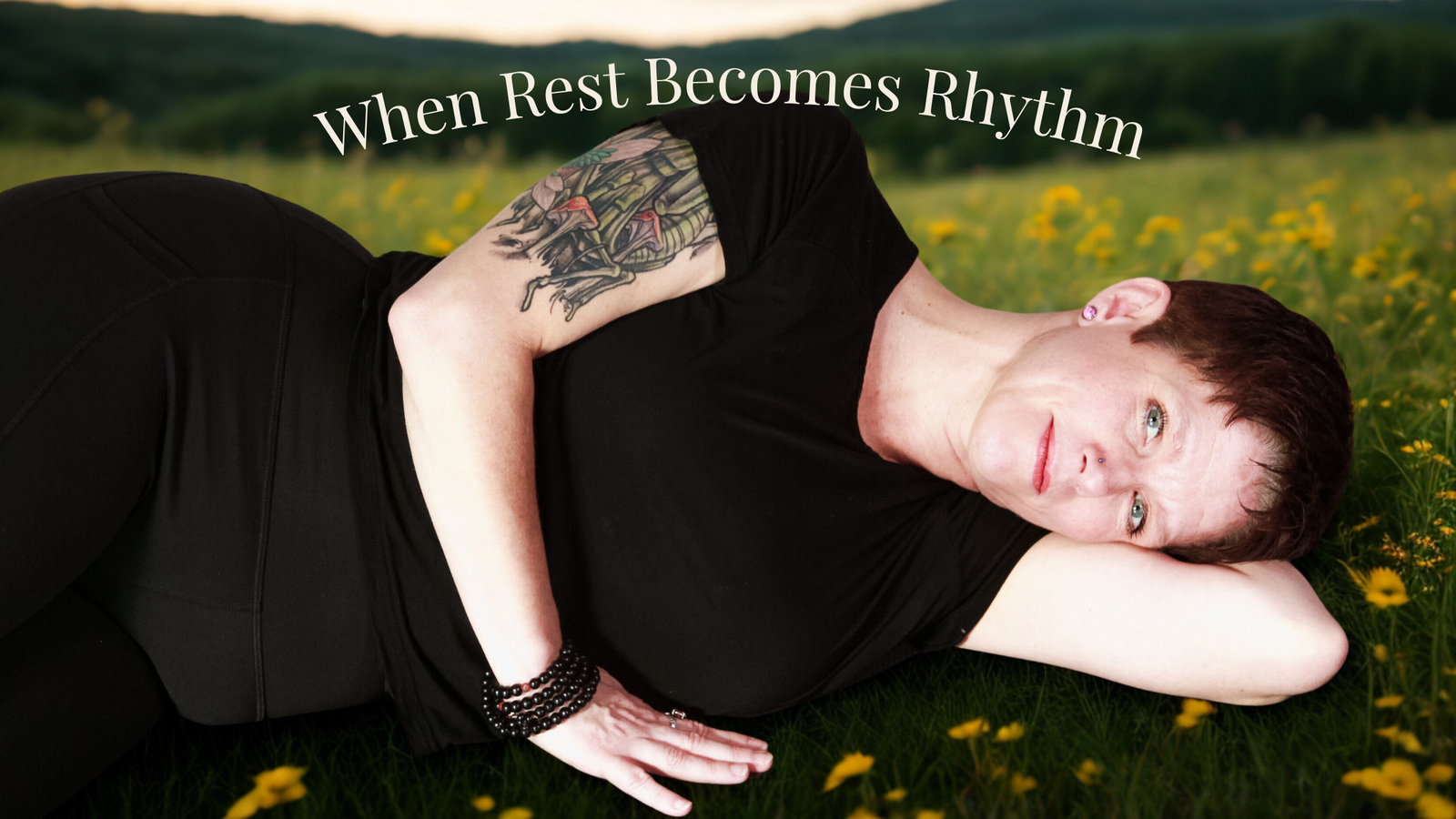 Yoga teacher resting in a side-lying position on grass in a field of yellow flowers, eyes open in quiet awareness, with the words “When Rest Becomes Rhythm” above.