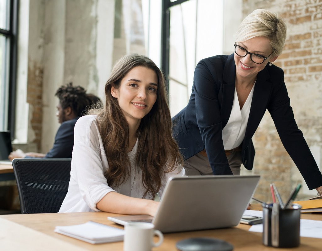 confident employee smiling at desk with laptop, mentored by experienced coworker in background