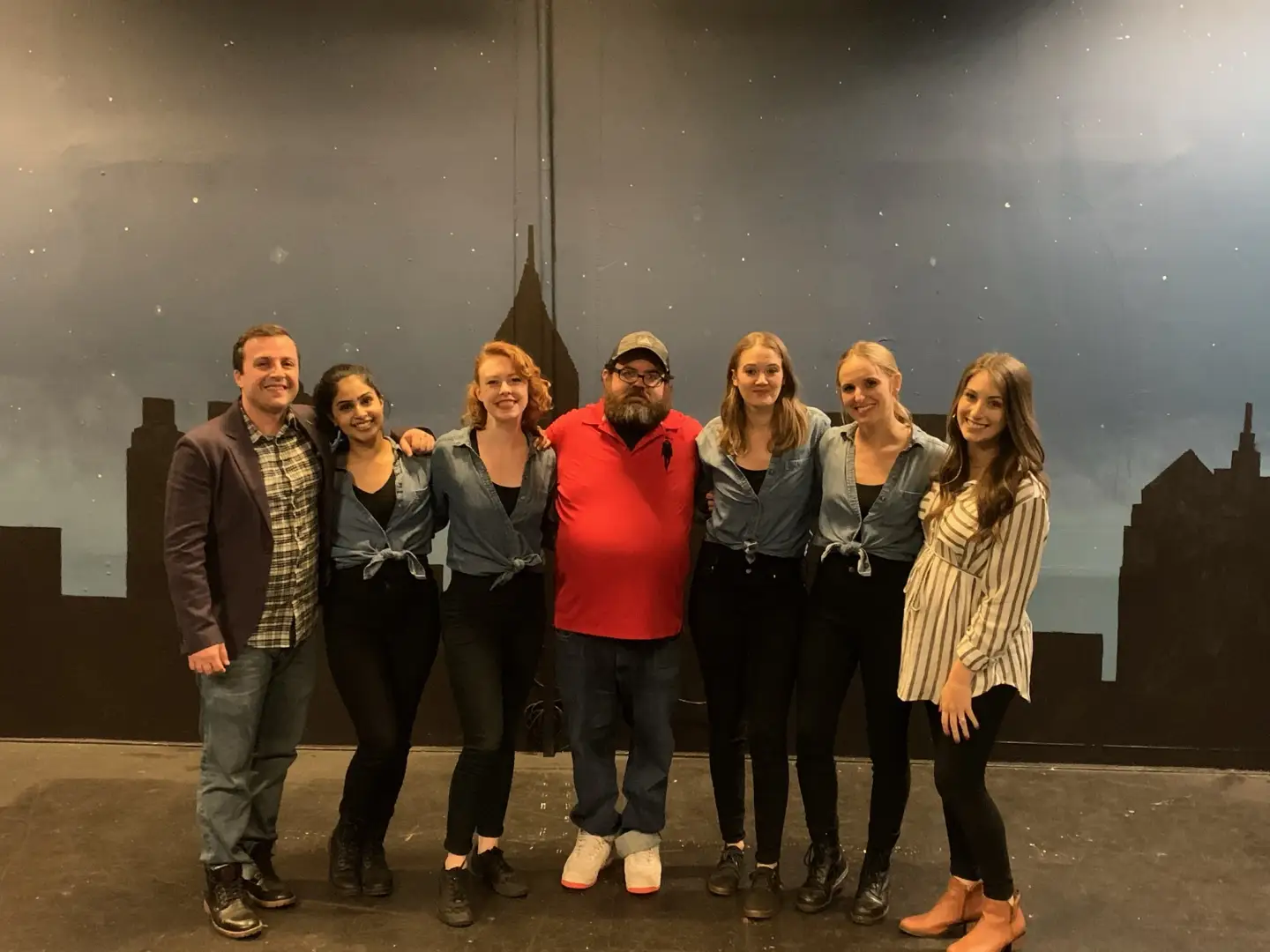 A group of seven cast and crew members pose together in front of a painted city-skyline backdrop.