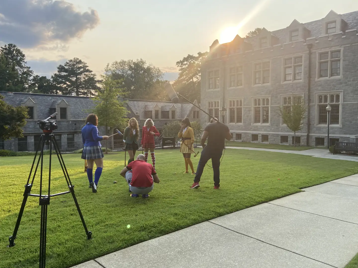 A film crew with a boom microphone shoots a scene with actors in school uniforms on a lawn at sunset, in front of a stone campus building.