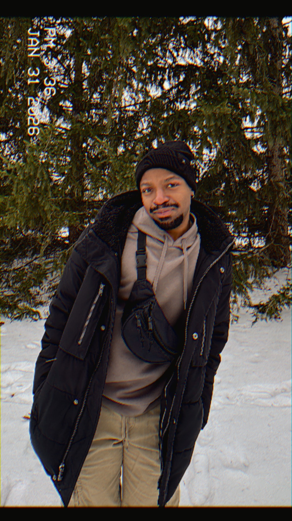 A man poses in a winter setting, wearing a stylish black jacket and beanie. The photo has a vintage film aesthetic.