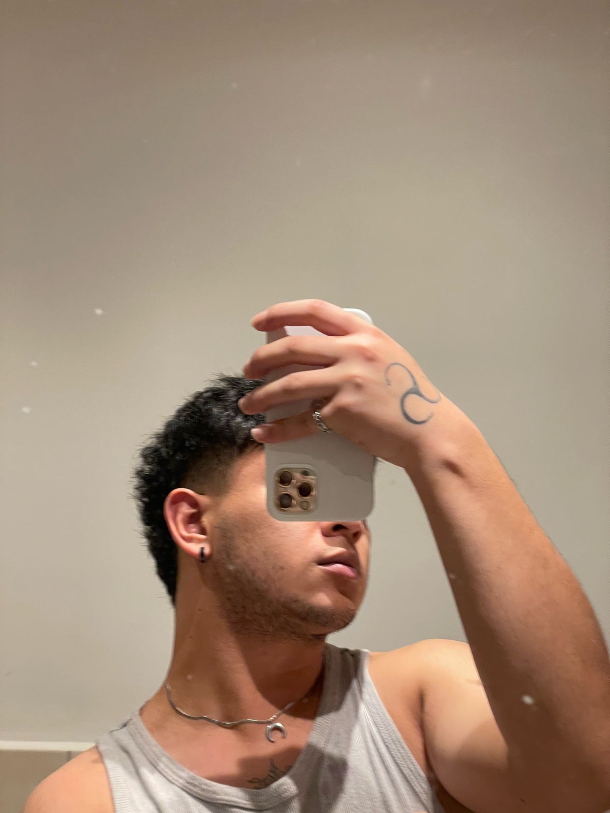 A man takes a mirror selfie, showcasing his curly hair and a tattoo on his hand. He is shirtless, wearing a tank top and jewelry.