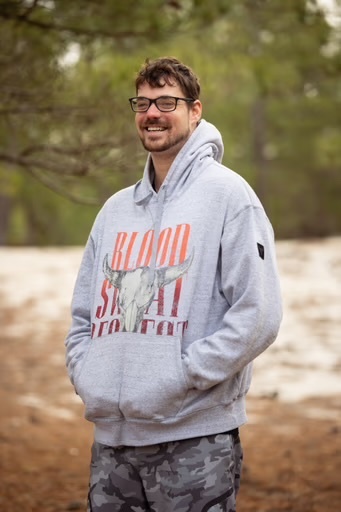 A man stands outdoors, smiling at the camera. He wears a grey hoodie with a graphic on the front.