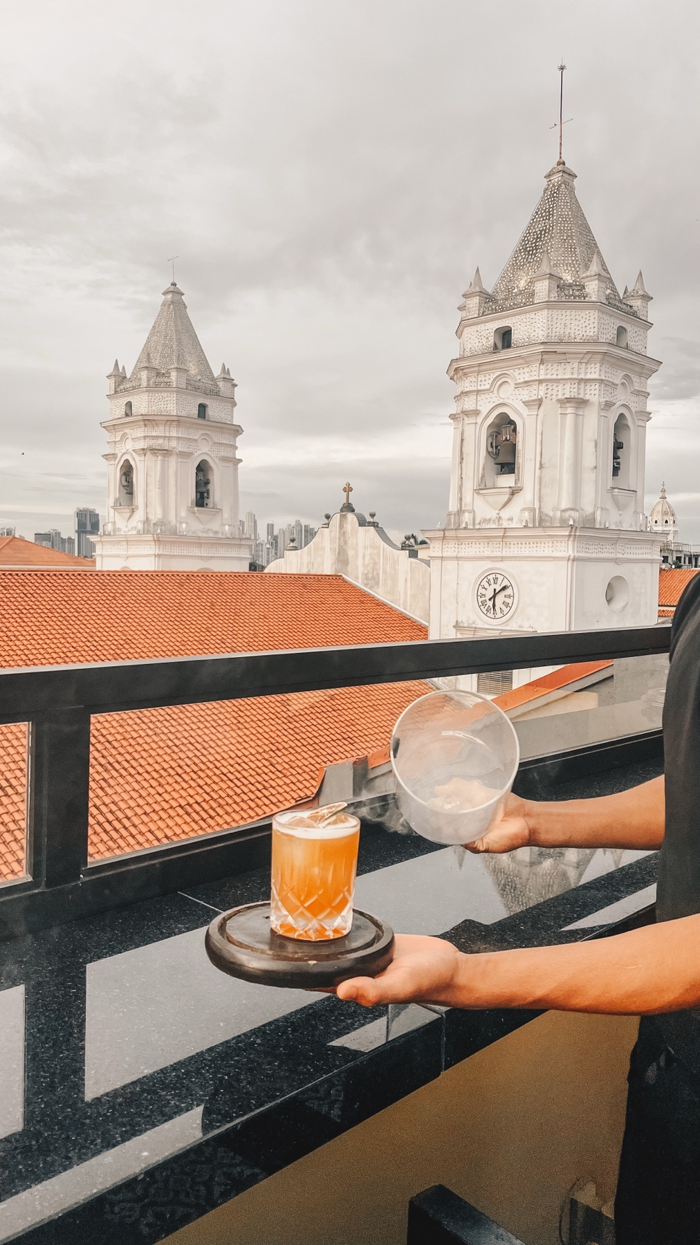 A person is preparing a smoky cocktail on a rooftop bar with a view of two white bell towers and a cityscape in the background.