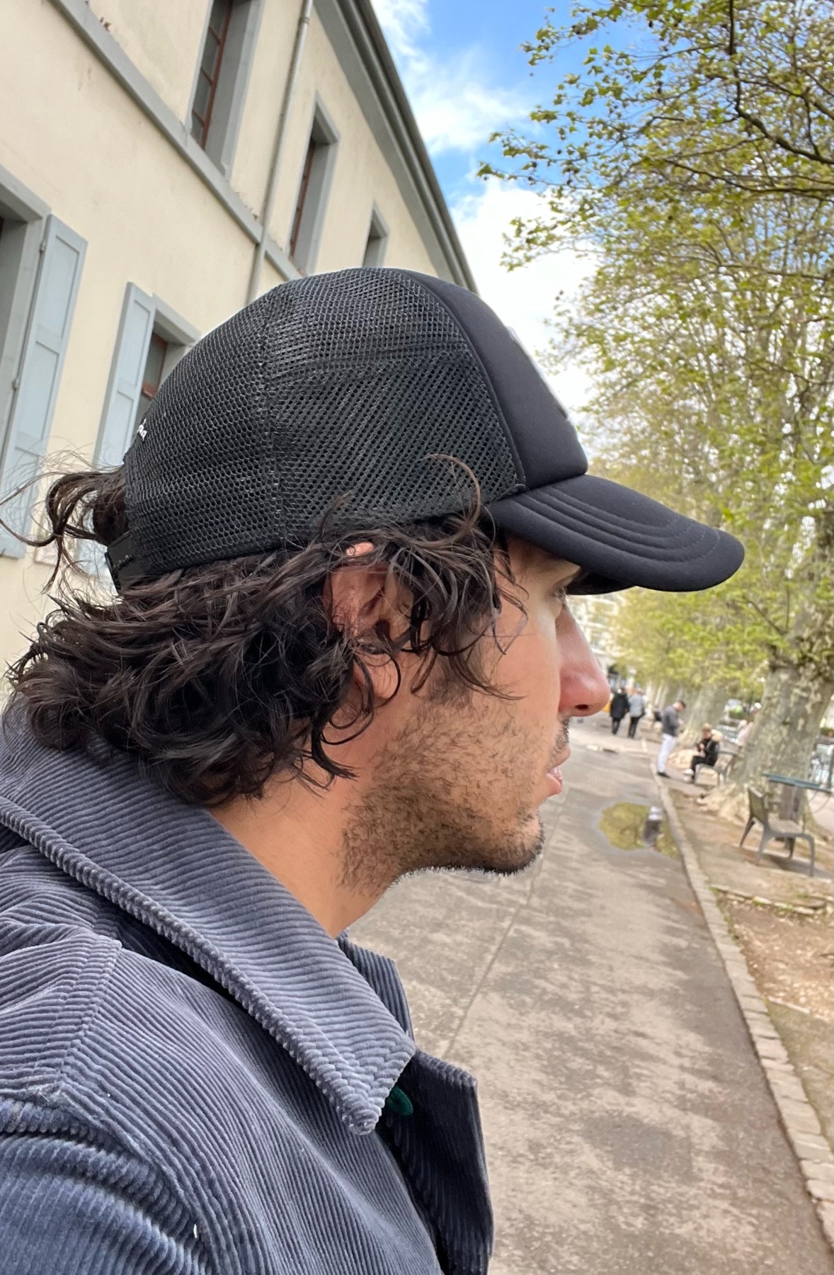 A man wearing a black trucker hat and a corduroy jacket is shown in profile, looking to the right. His curly hair is visible beneath the hat, and he has stubble on his face. The background shows a building and a tree-lined walkway.