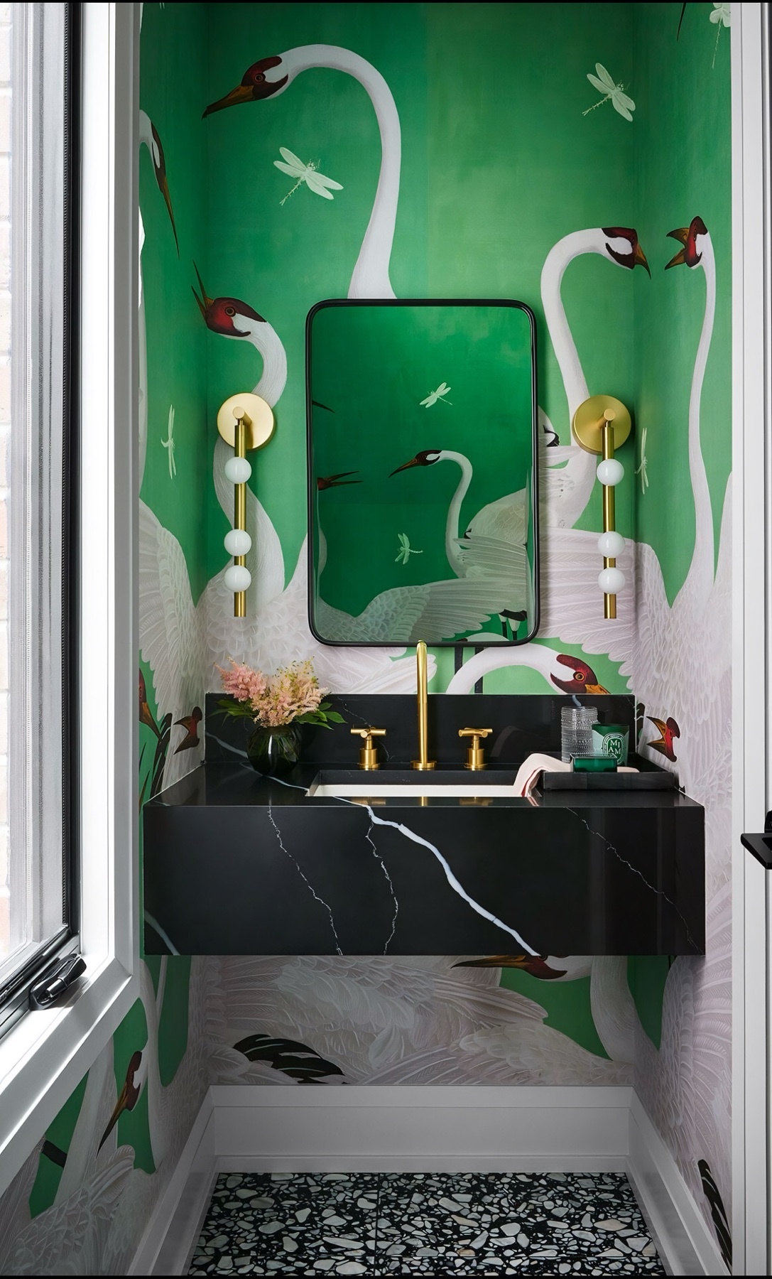 A stylish bathroom features bold green wallpaper with a crane and dragonfly pattern. A sleek black marble vanity with gold fixtures sits beneath a matching mirror, flanked by modern sconces.