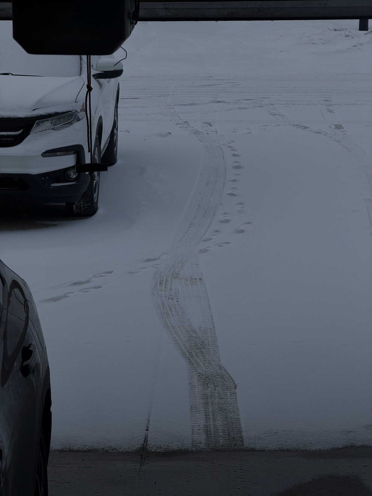 Tire tracks and footprints are visible in the snow on a parking lot, with the front of a white car on the left.