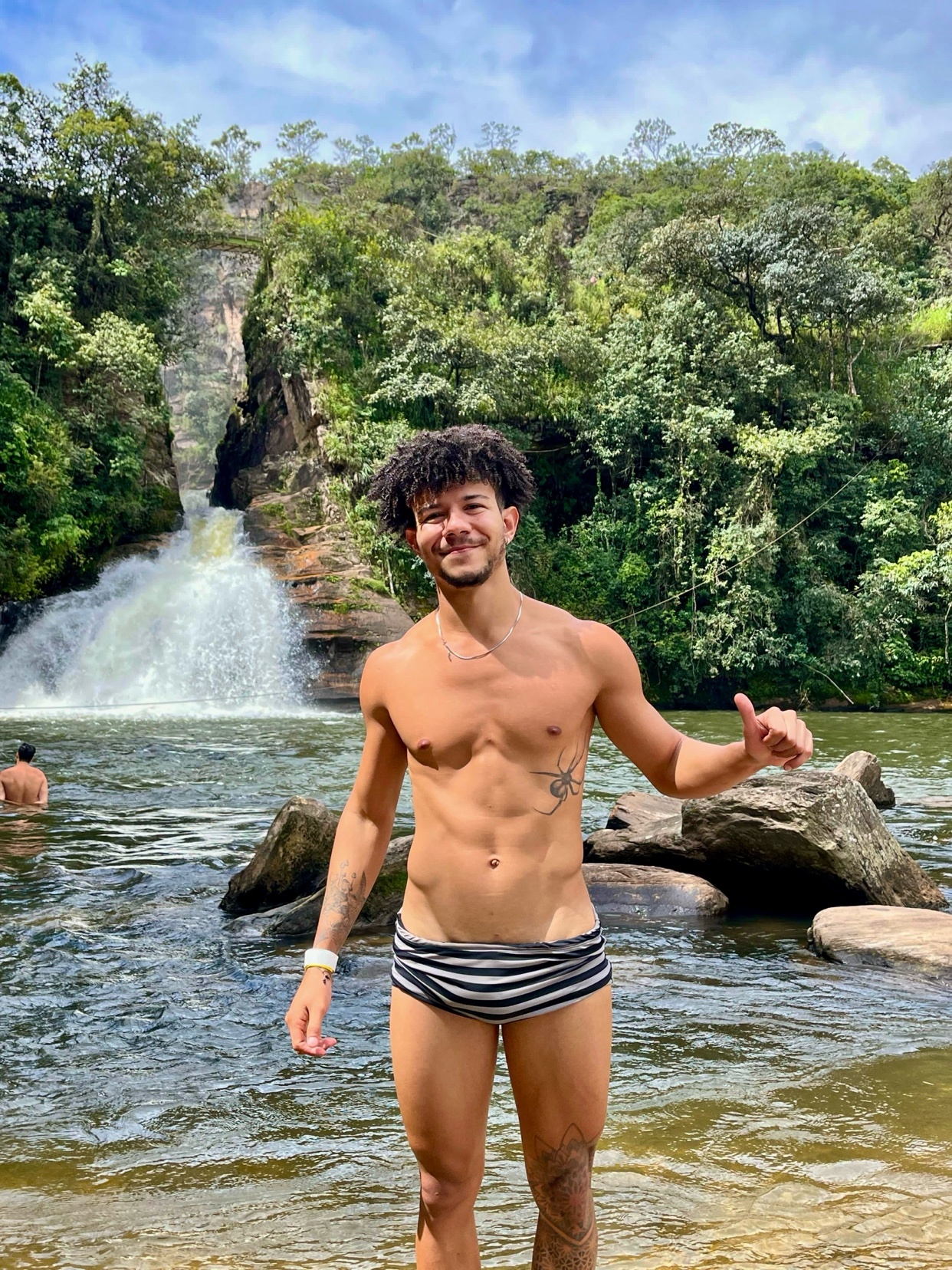 A man with curly hair and tattoos poses shirtless in striped swimwear by a waterfall, giving a thumbs up.
