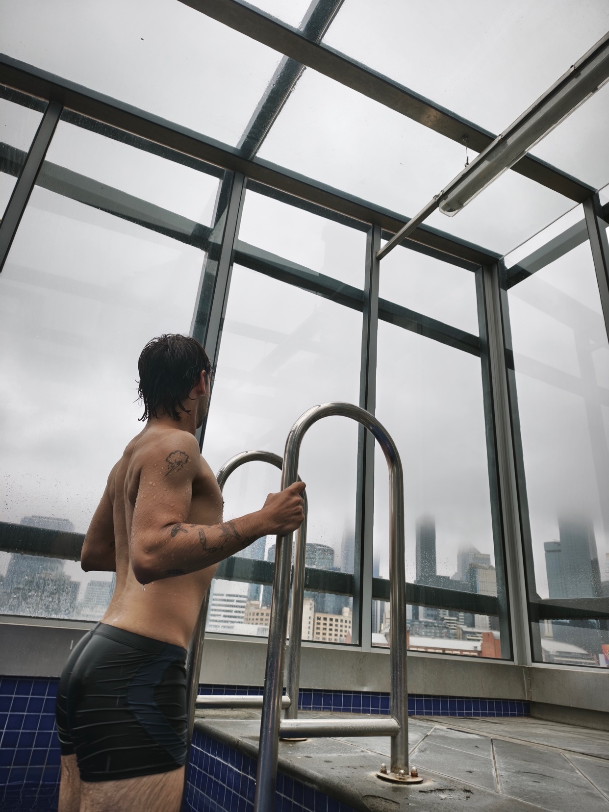 A man stands shirtless in a pool, looking out at a cityscape through a large window. The photo captures a rainy day atmosphere.