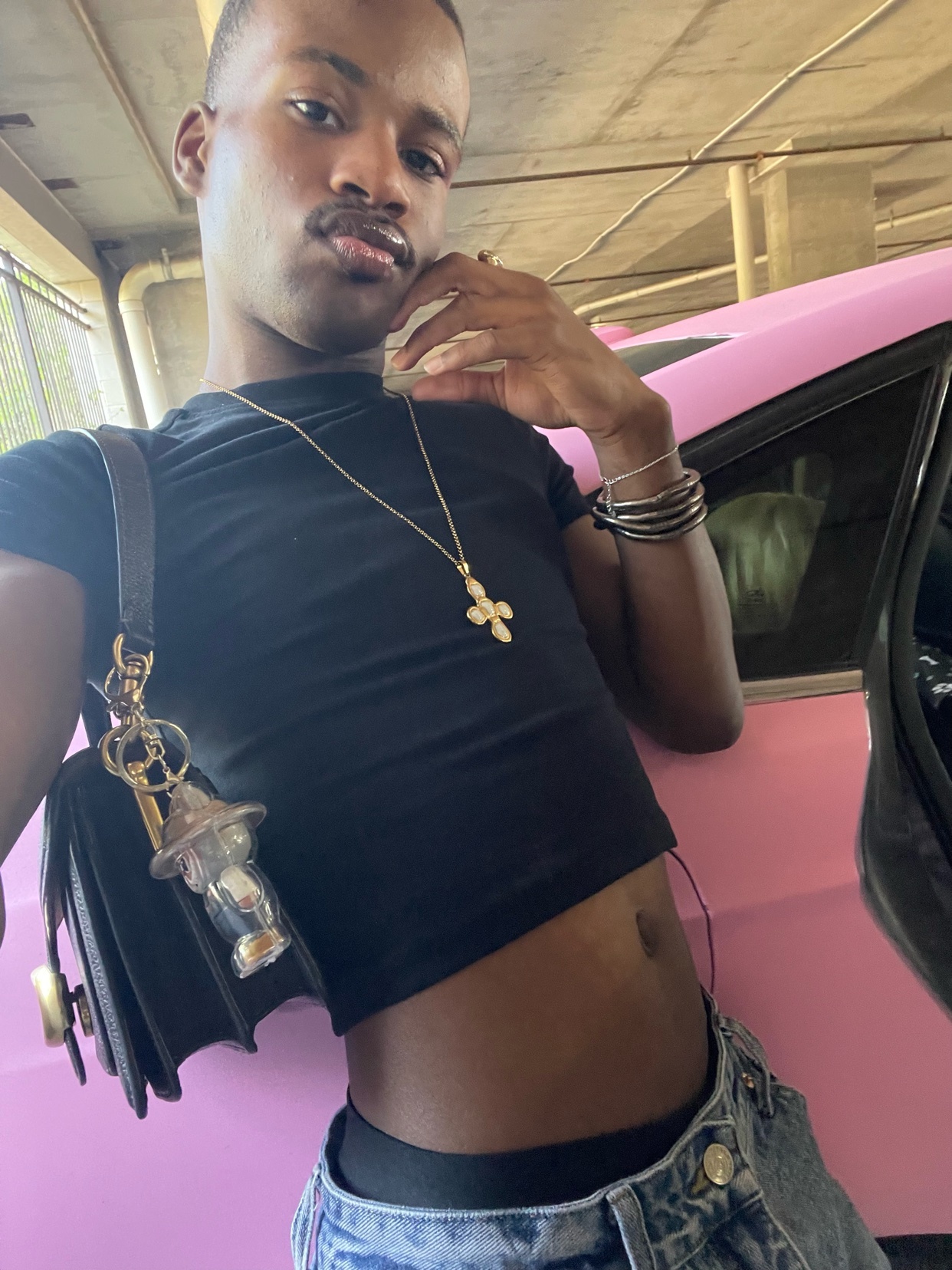 A man in a black cropped t-shirt and denim jeans poses for a selfie next to a pink car. He is wearing a cross necklace and holding a handbag.