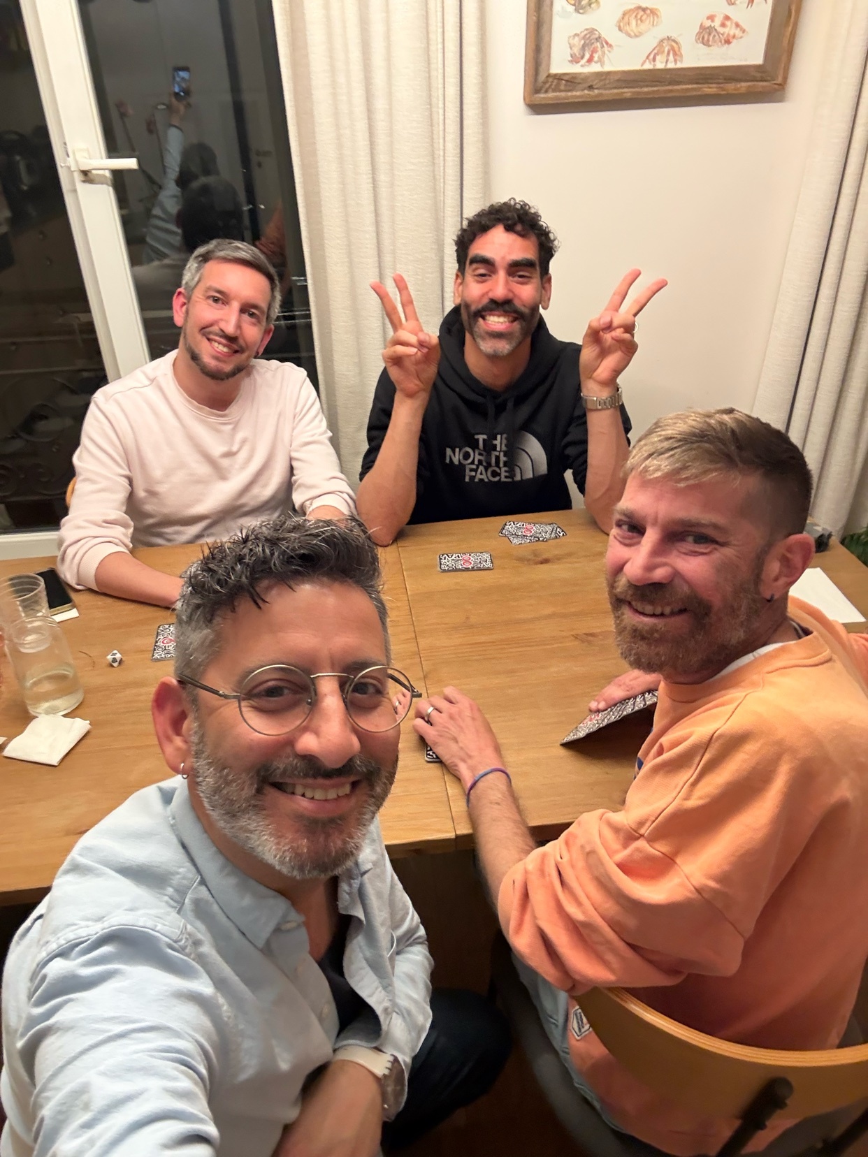 Four men are gathered around a wooden table, playing a card game. One man in the foreground is taking a selfie, while another in the background flashes a peace sign.