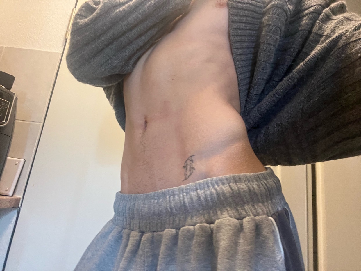 A close-up, low-angle shot of a person's torso, showing their abs and a small tattoo on their hip. They are wearing grey sweatpants and a grey cardigan draped over their shoulders.