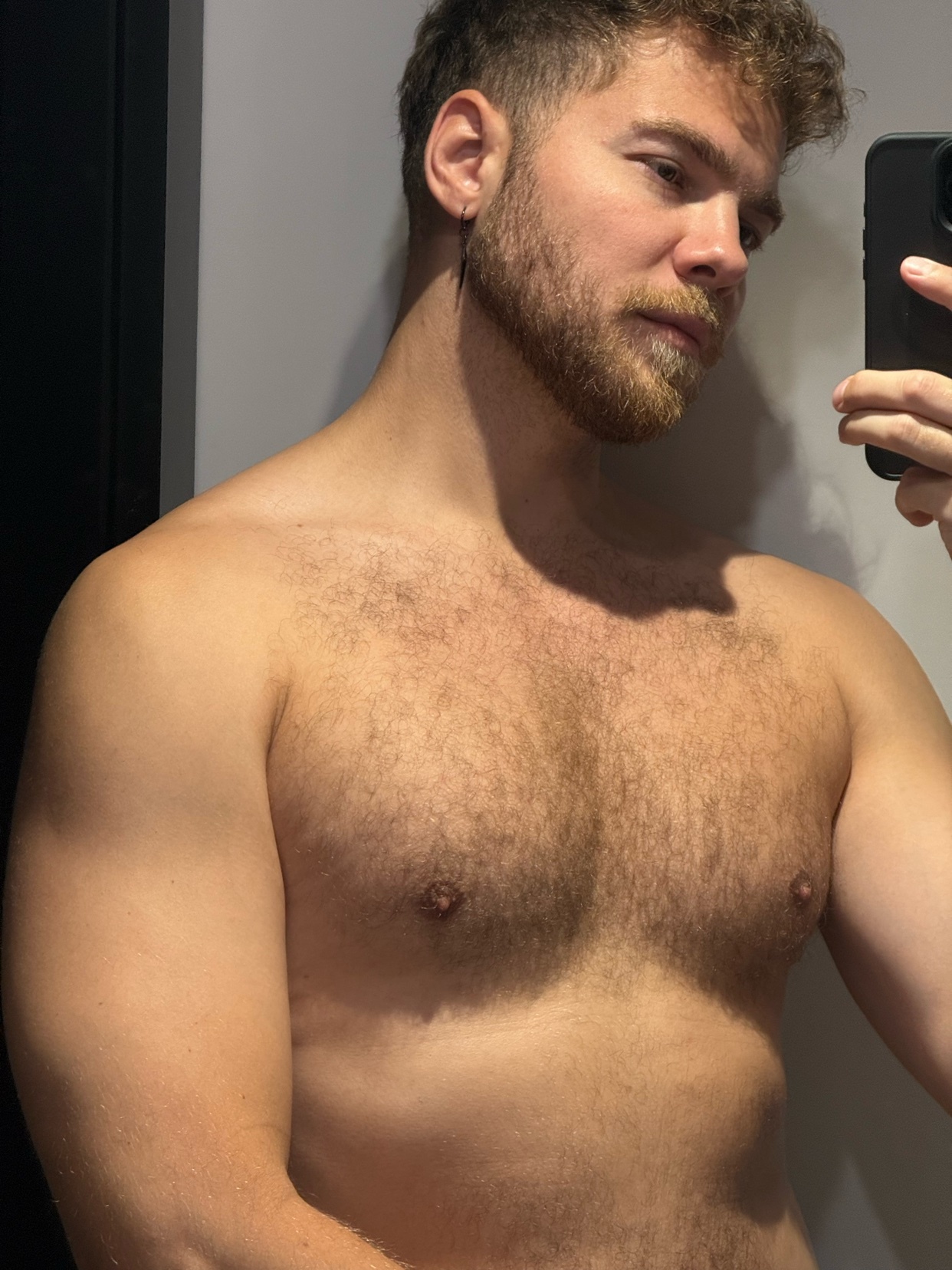 A man with curly hair and a beard takes a mirror selfie. He is shirtless, showing his chest hair and muscular arms.