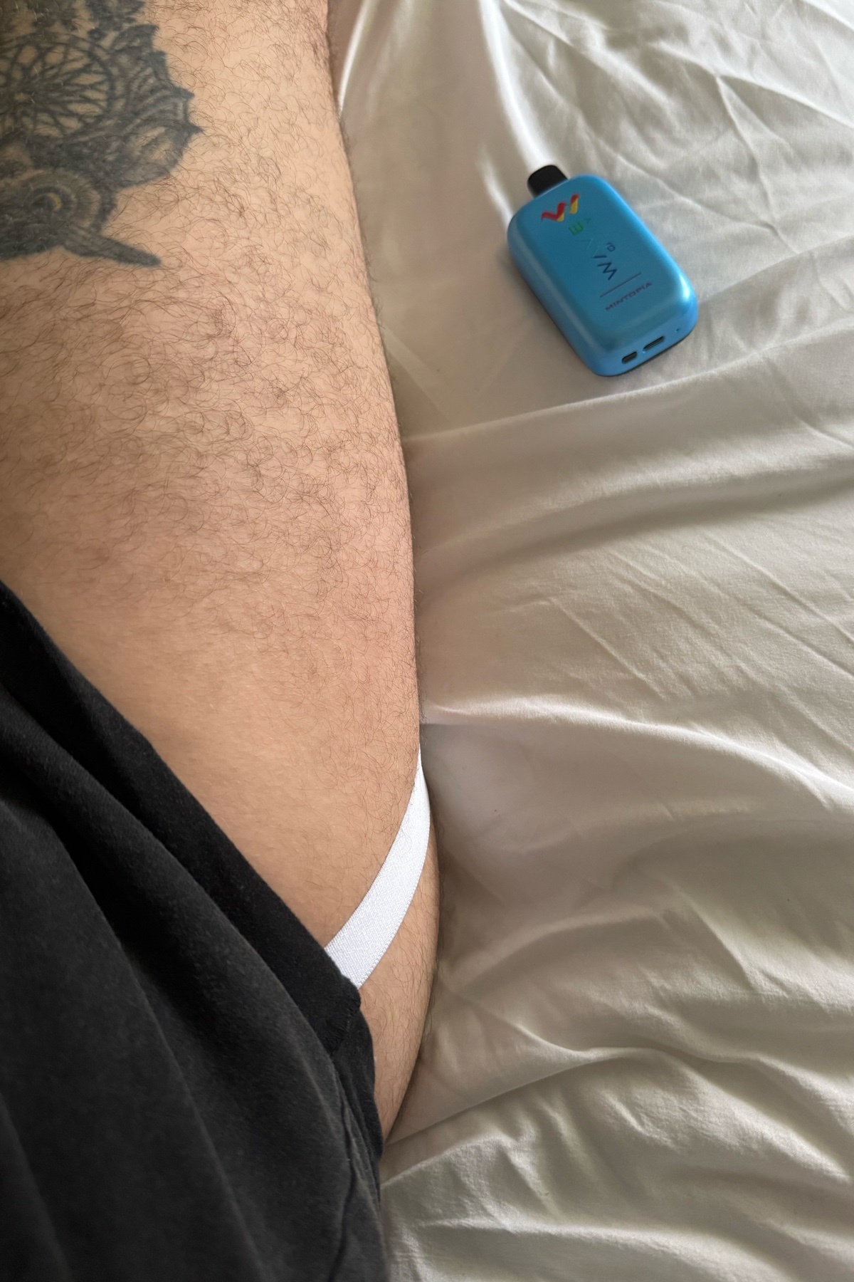 A man's leg is visible, wearing a white jockstrap. A vape device sits on the bed next to him.
