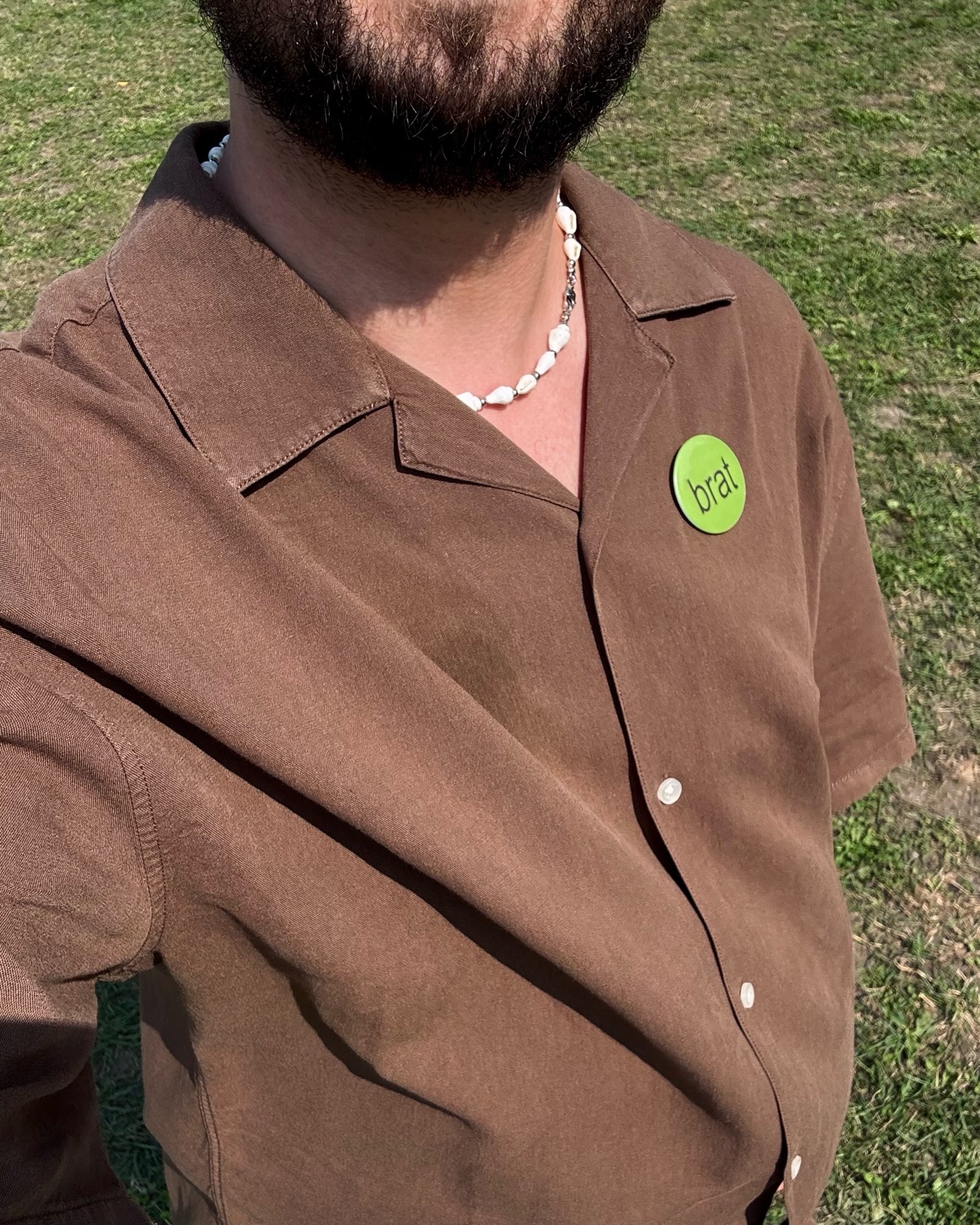 A man takes a selfie, wearing a brown button-up shirt and a pearl necklace. The photo is taken outside on a sunny day.
