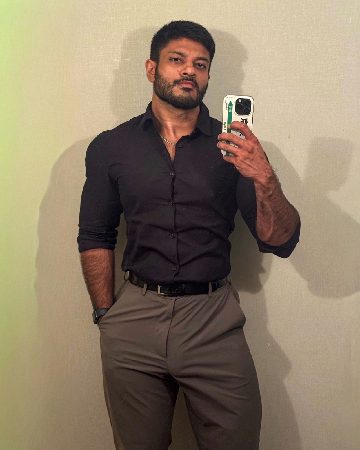 A man poses for a selfie, wearing a black button-down shirt and grey pants. He has a beard and muscular physique.