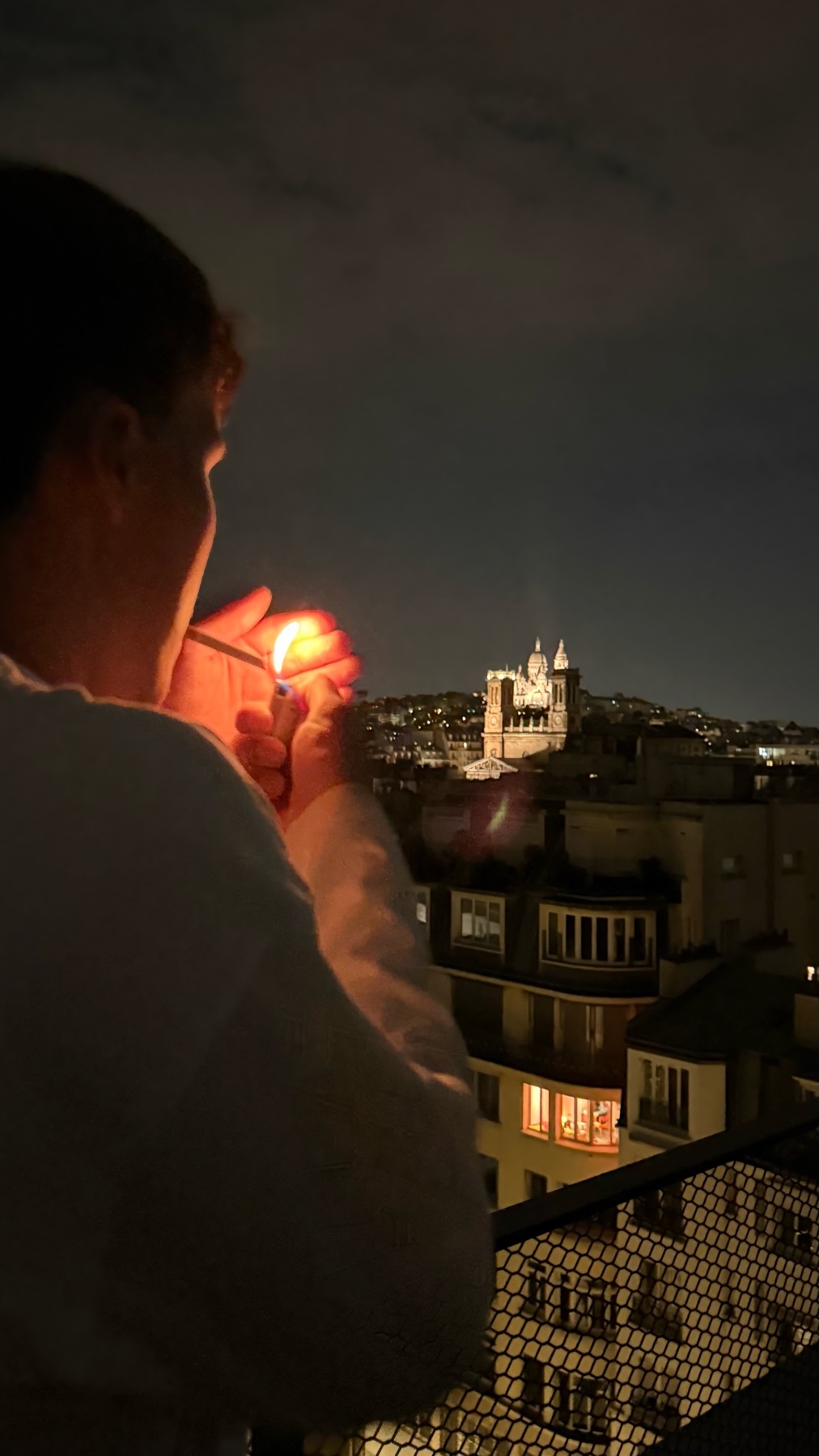 A person lights a cigarette on a balcony at night with a city skyline in the background.
