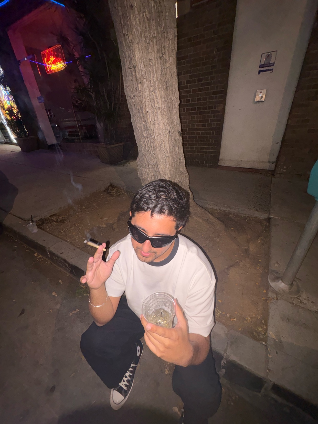 A man wearing sunglasses and a t-shirt squats on a sidewalk at night, holding a drink and smoking a cigarette.