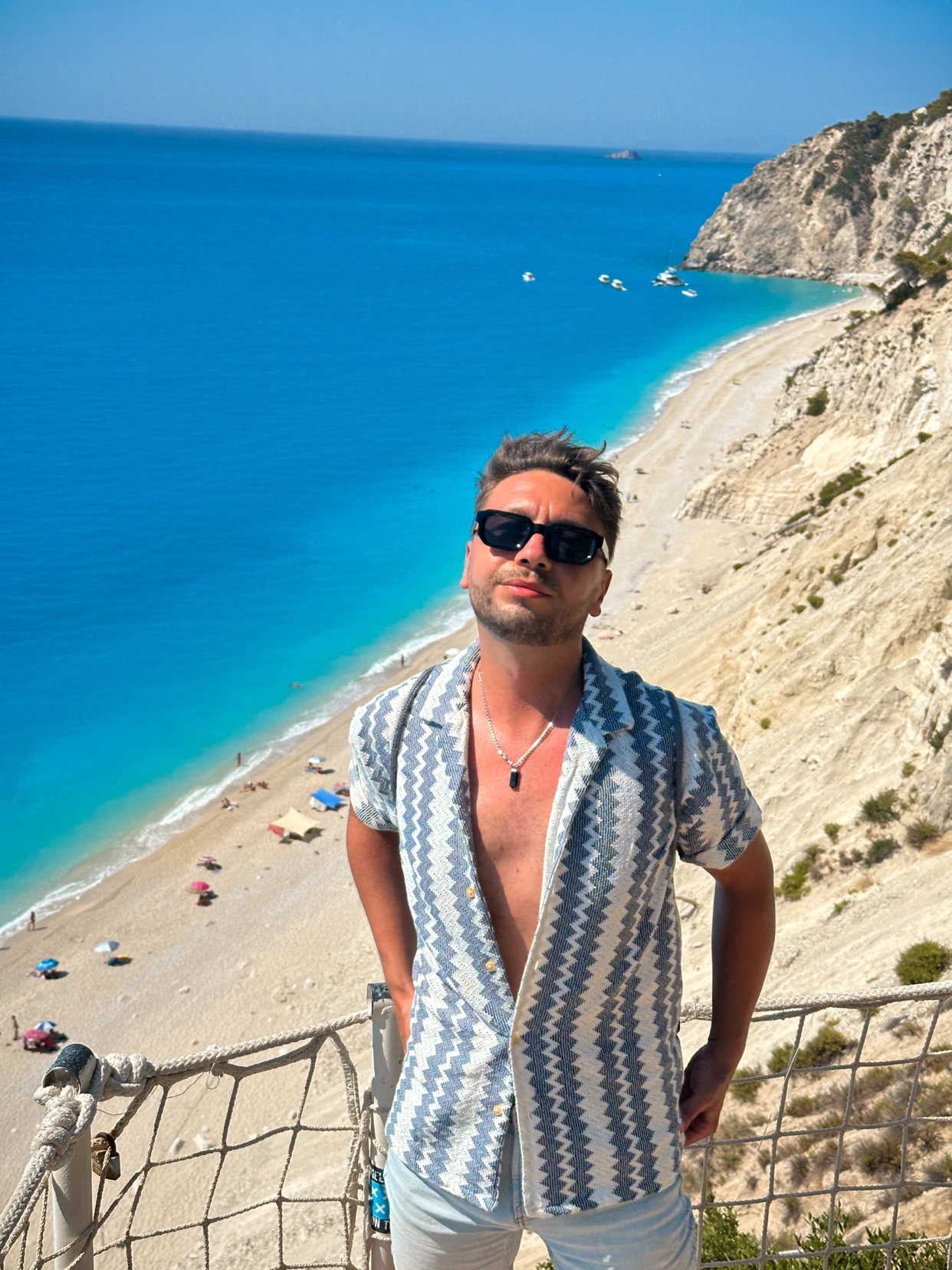 A man poses on a cliff overlooking a beautiful beach and ocean. He is wearing a stylish shirt and sunglasses.