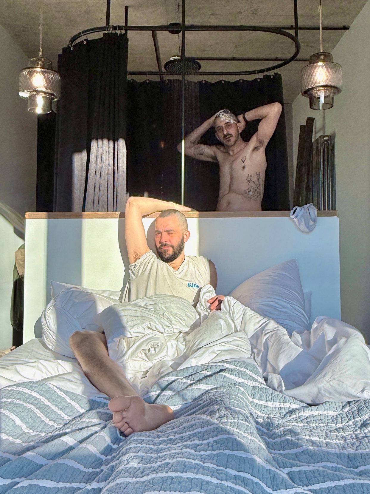 Two men are relaxing in bed in the morning light. One man is lying in bed under the covers, while the other is standing behind him, leaning against the headboard.