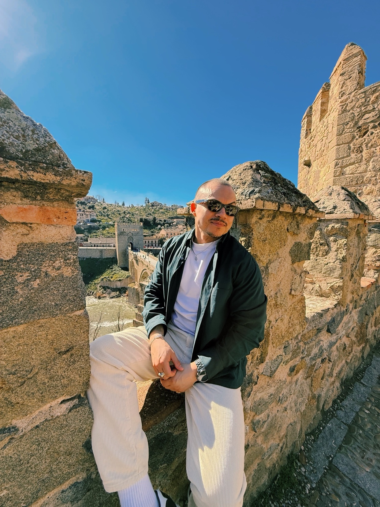 A man with a bald head and stubble poses while sitting on a stone wall with a clear blue sky above. He is wearing sunglasses, a white t-shirt, a dark jacket, and light-colored pants.
