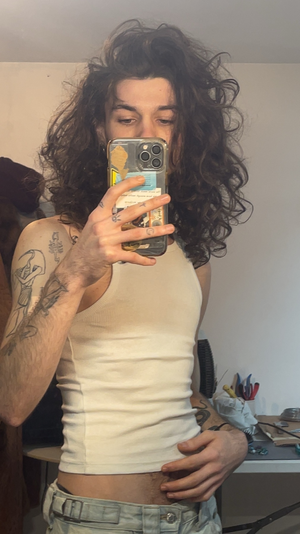 A man takes a mirror selfie, showcasing his long, curly hair and tattoos. He is shirtless, wearing a tank top.