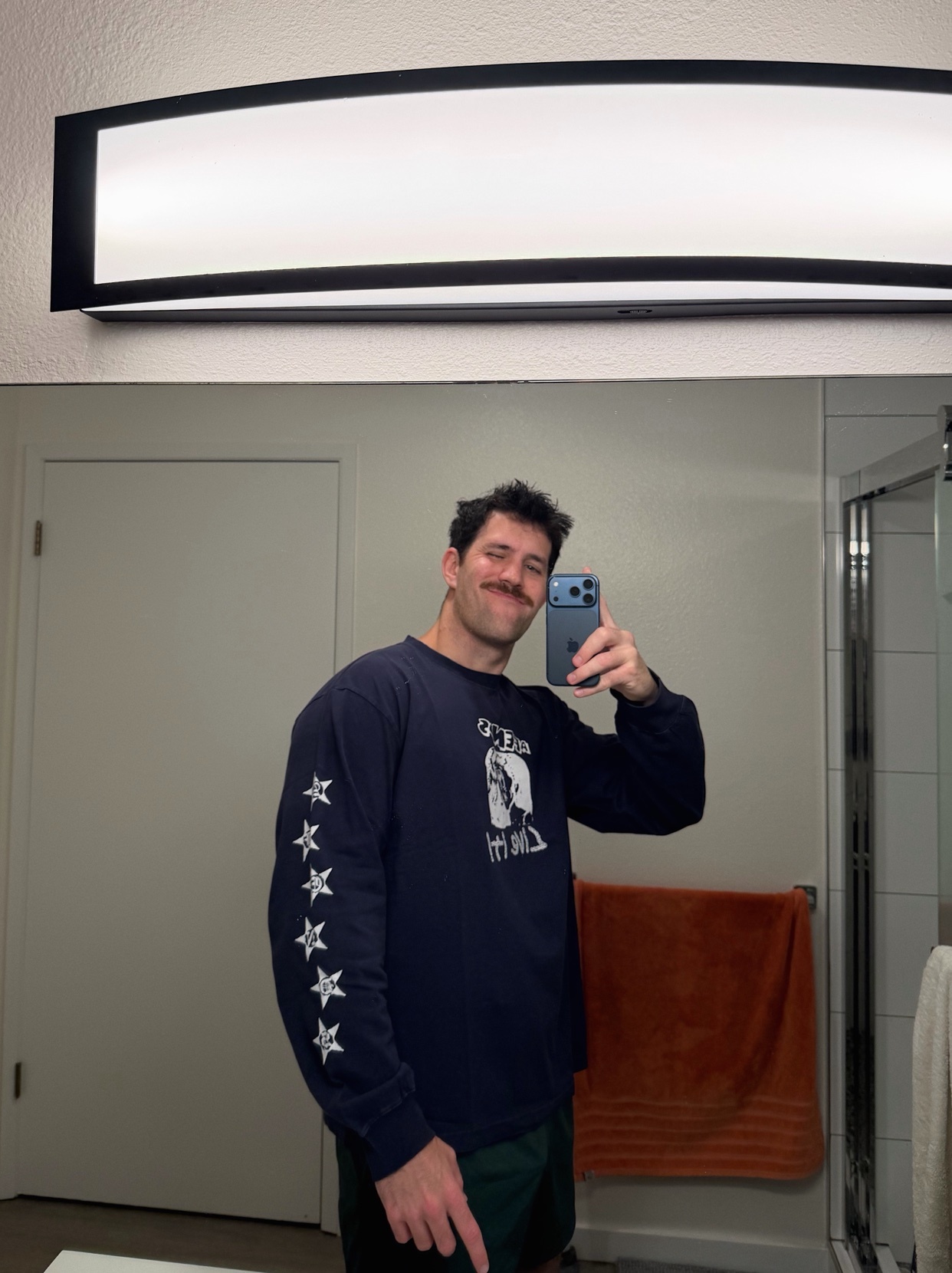 A man with a mustache takes a mirror selfie in a bathroom. He is wearing a navy blue long-sleeved shirt with white stars on the sleeve and dark green shorts.