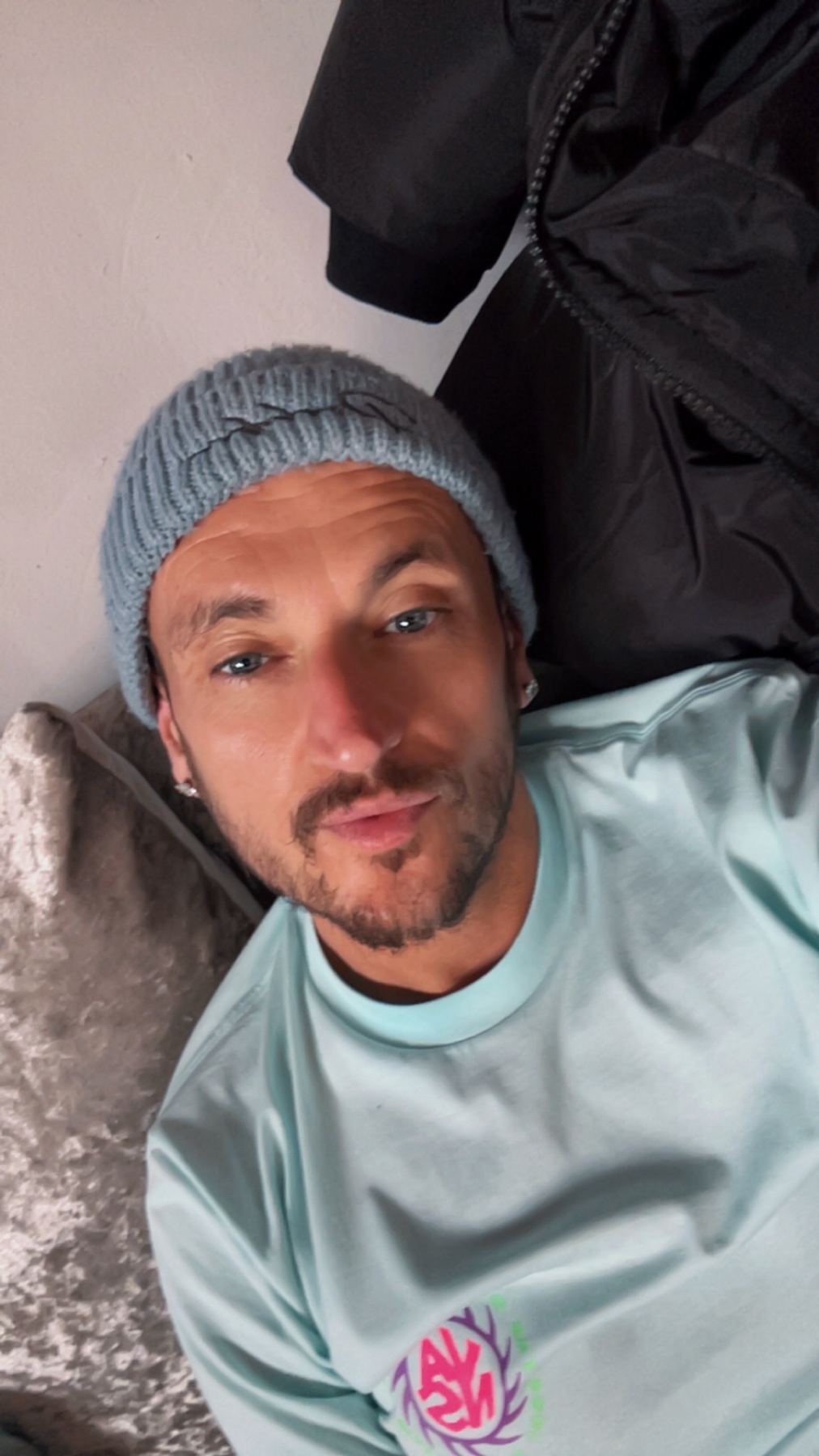 A man with stubble and blue eyes wears a light blue beanie and a light blue t-shirt. He is looking at the camera in a selfie.