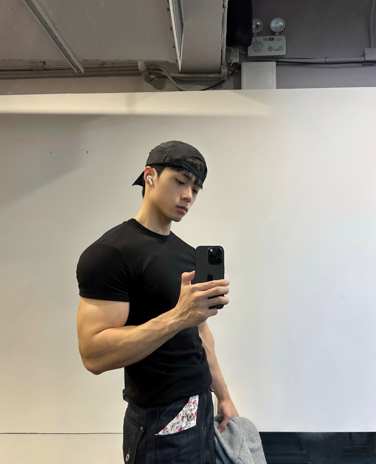 A man takes a mirror selfie at the gym. He is wearing a black t-shirt and a cap, showing off his muscular physique.