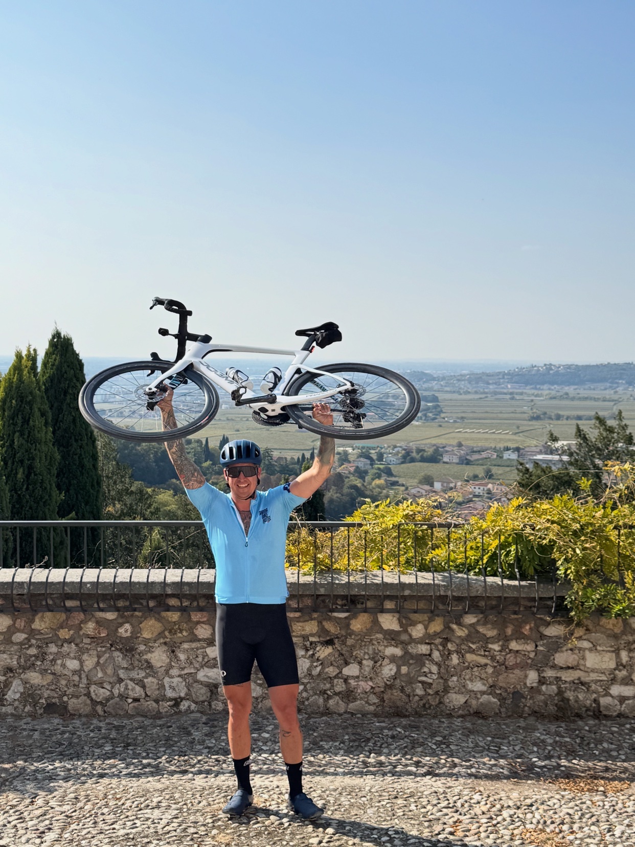 A man in cycling attire holds a bicycle above his head in a triumphant pose. He is outdoors with a scenic view of rolling hills and a town in the distance.