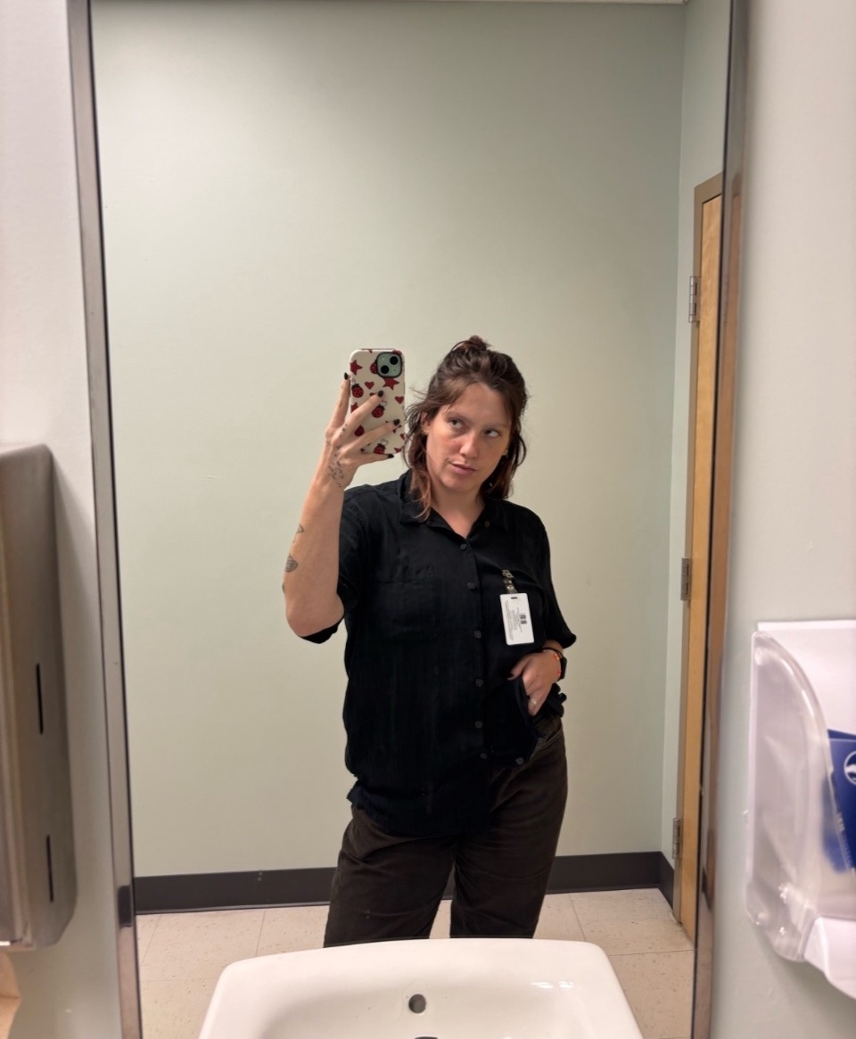 A woman takes a mirror selfie in a bathroom, wearing a black button-down shirt and dark pants. She has an ID badge clipped to her shirt.