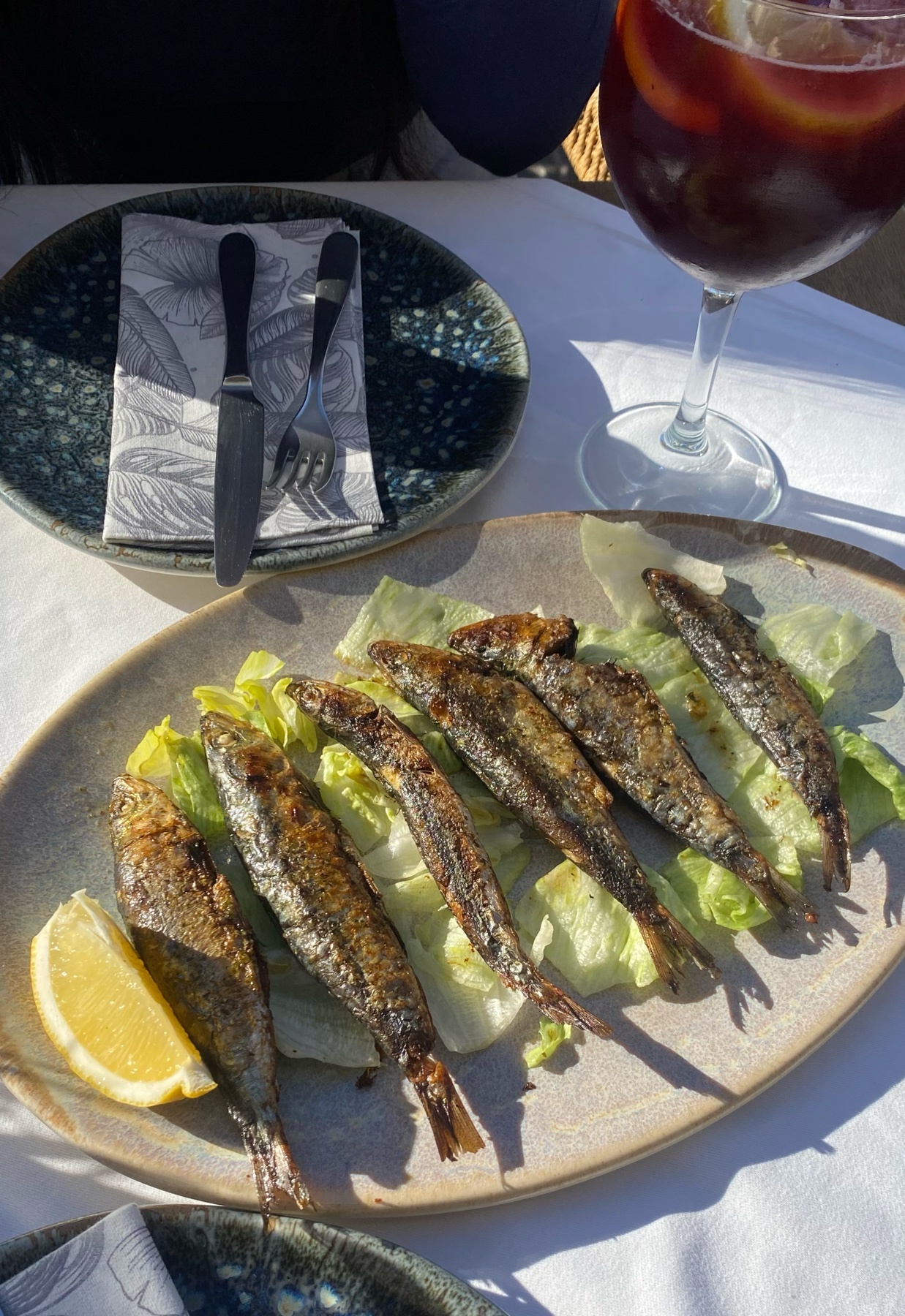 A plate of fried sardines is presented with a lemon wedge and lettuce. A glass of red wine sits nearby on a white tablecloth.