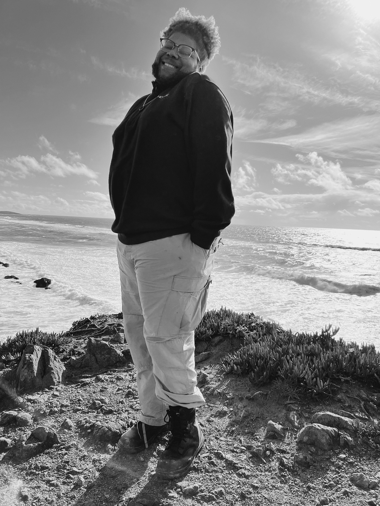 A man stands on a cliff overlooking the ocean. He is smiling and wearing casual clothing.