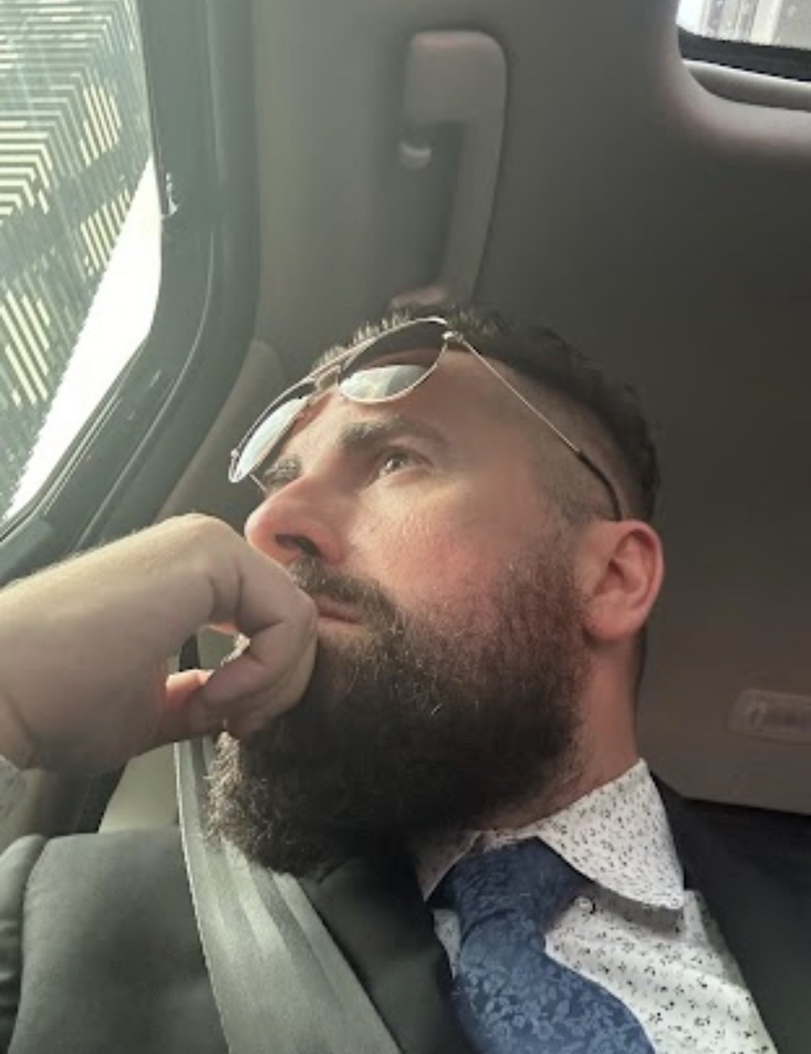 A man with a full beard and short undercut hair is wearing sunglasses, a suit, and a patterned tie. He is looking upwards and to the side, appearing to be in deep thought while inside a vehicle.
