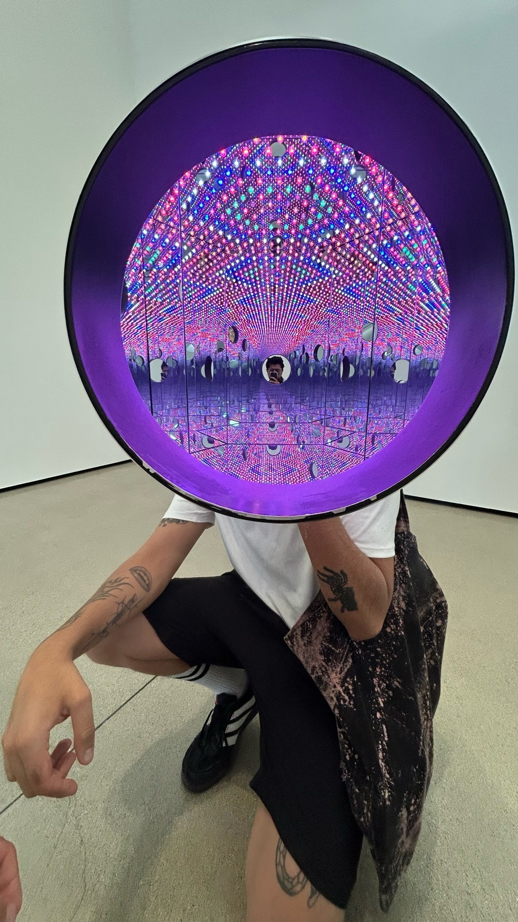 A person crouches in front of a large, purple-rimmed circular art installation that creates an infinite tunnel of lights. The installation reflects the surrounding gallery space and the person within it.
