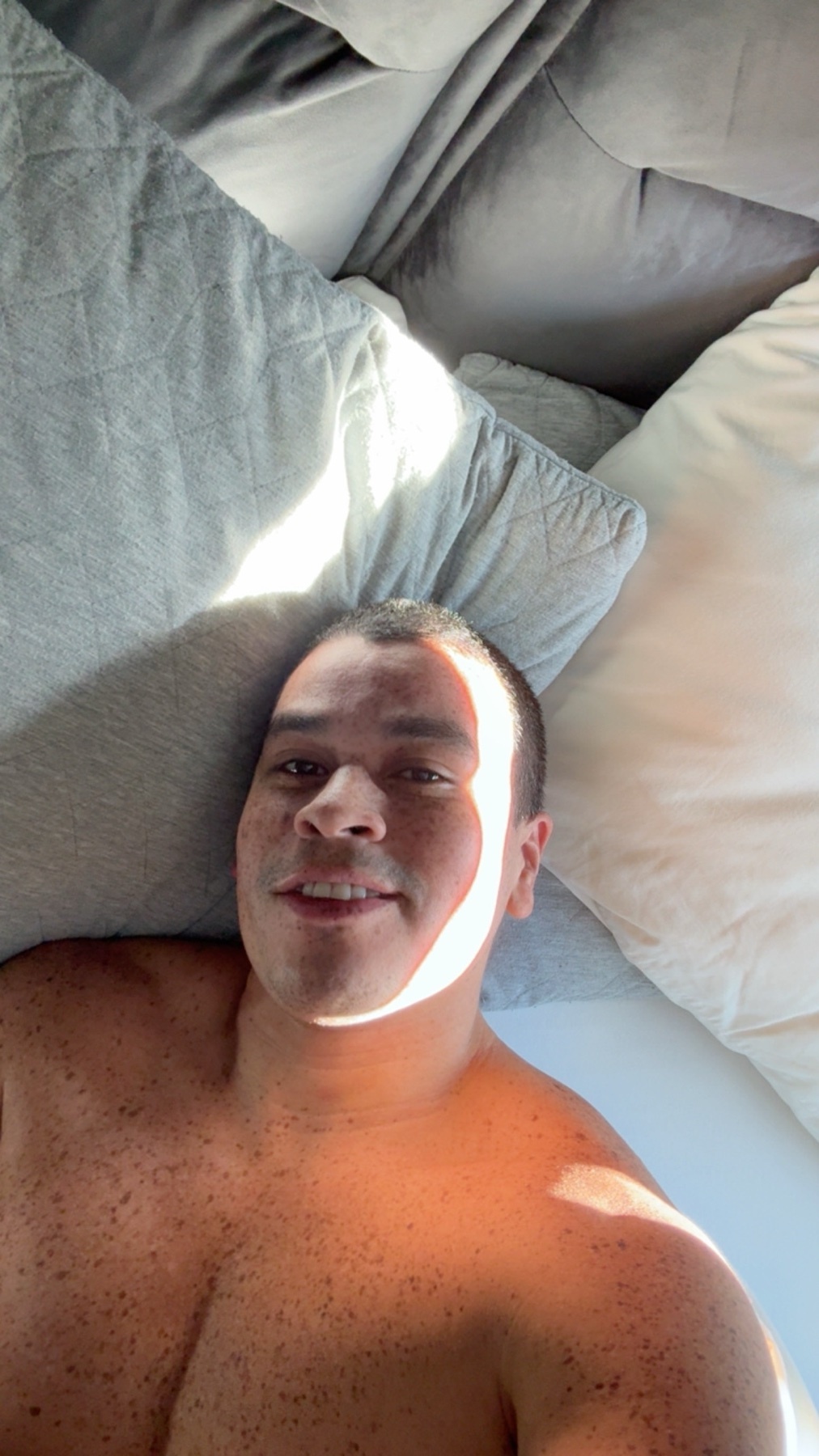 A shirtless man with freckles lies in bed, bathed in morning light. His muscular chest is visible as he smiles at the camera.