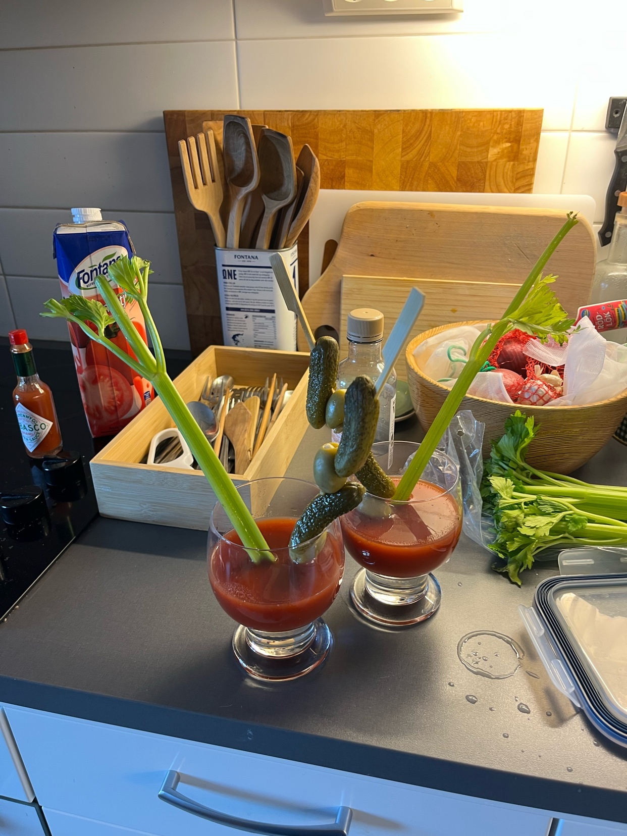 Two Bloody Mary cocktails garnished with celery and pickles sit on a kitchen counter next to a bottle of Tabasco sauce and a carton of tomato juice.