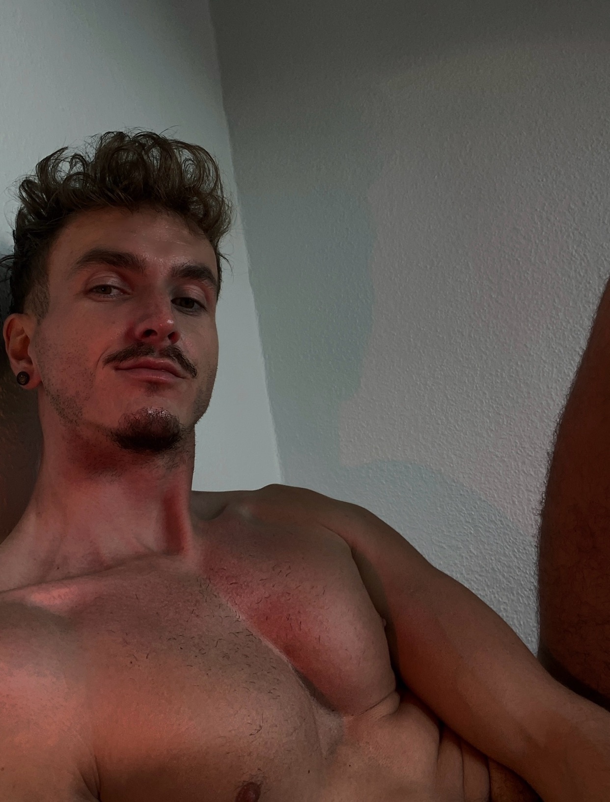 A man takes a selfie, shirtless, with curly hair and a mustache. The photo is taken from a low angle.