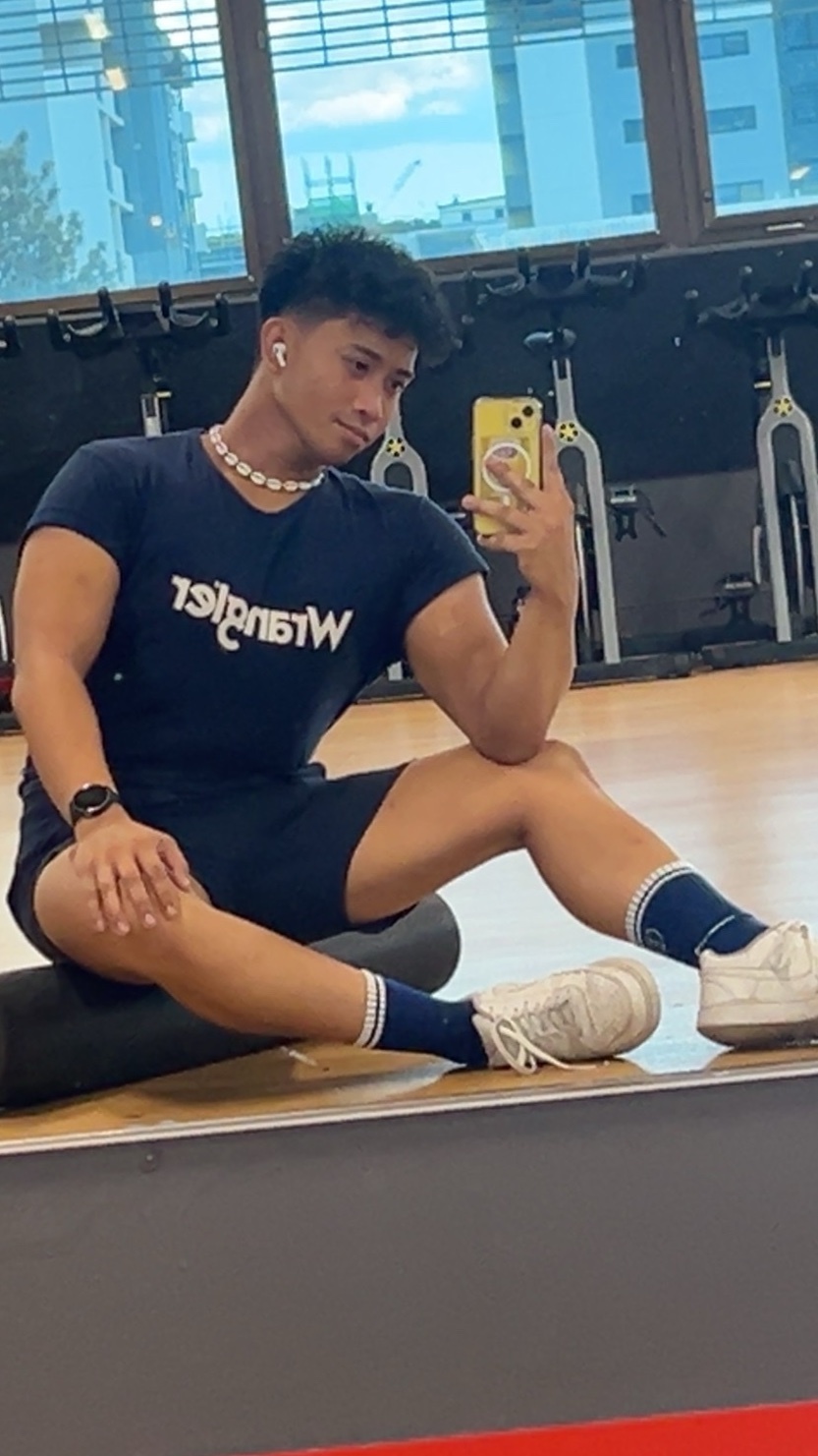 A young Asian man with tattoos on his arm poses for a mirror selfie in a gym. He is wearing a dark t-shirt, short shorts, crew socks, and sneakers, sitting on a foam roller.
