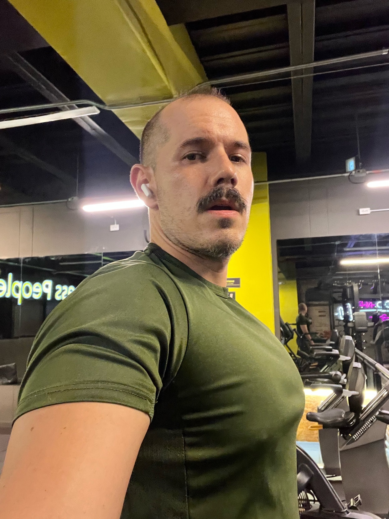 A man with a mustache poses for a selfie in a gym, wearing an olive green t-shirt.