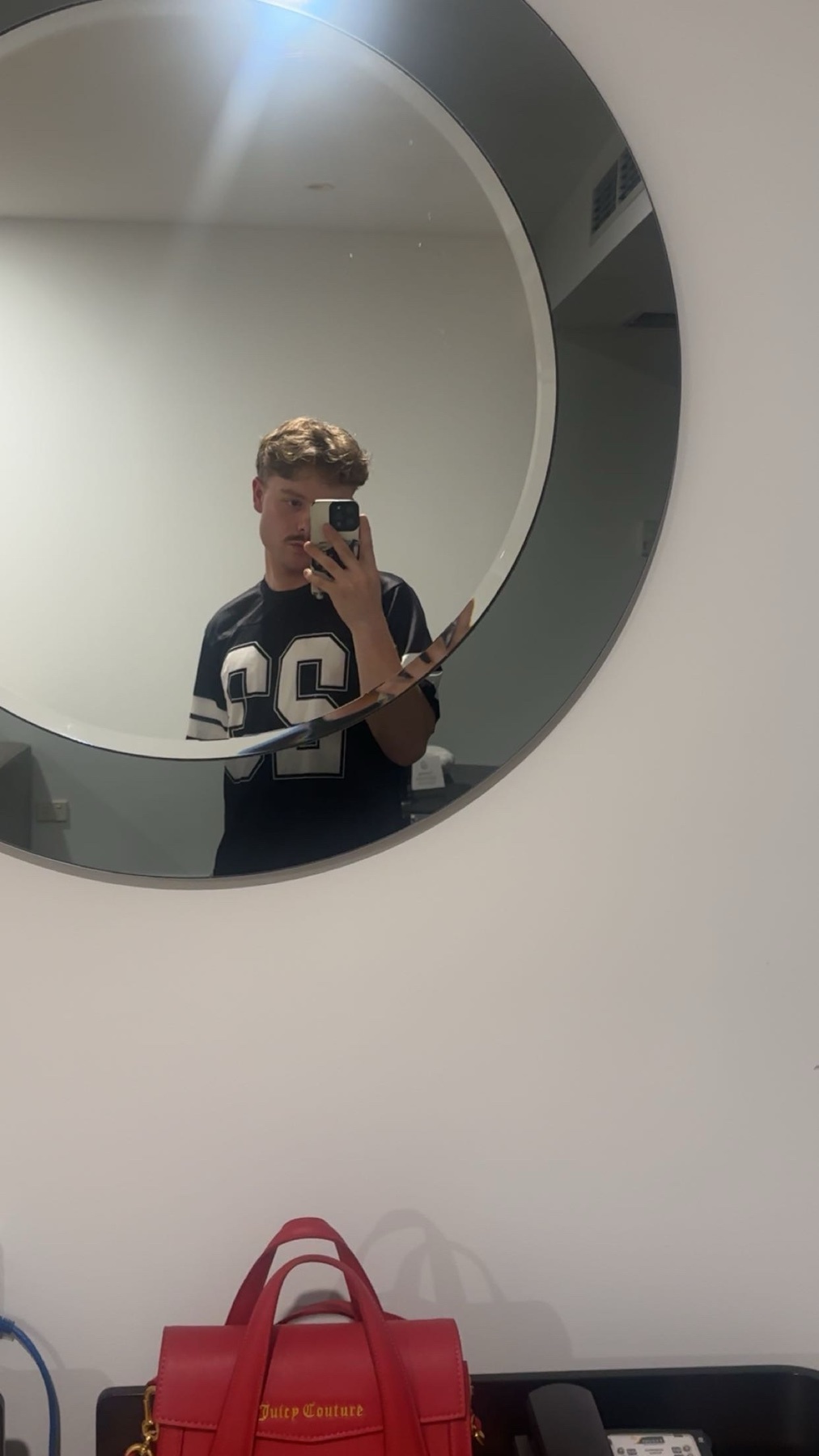 A man takes a mirror selfie. He is wearing a jersey and holding a phone.