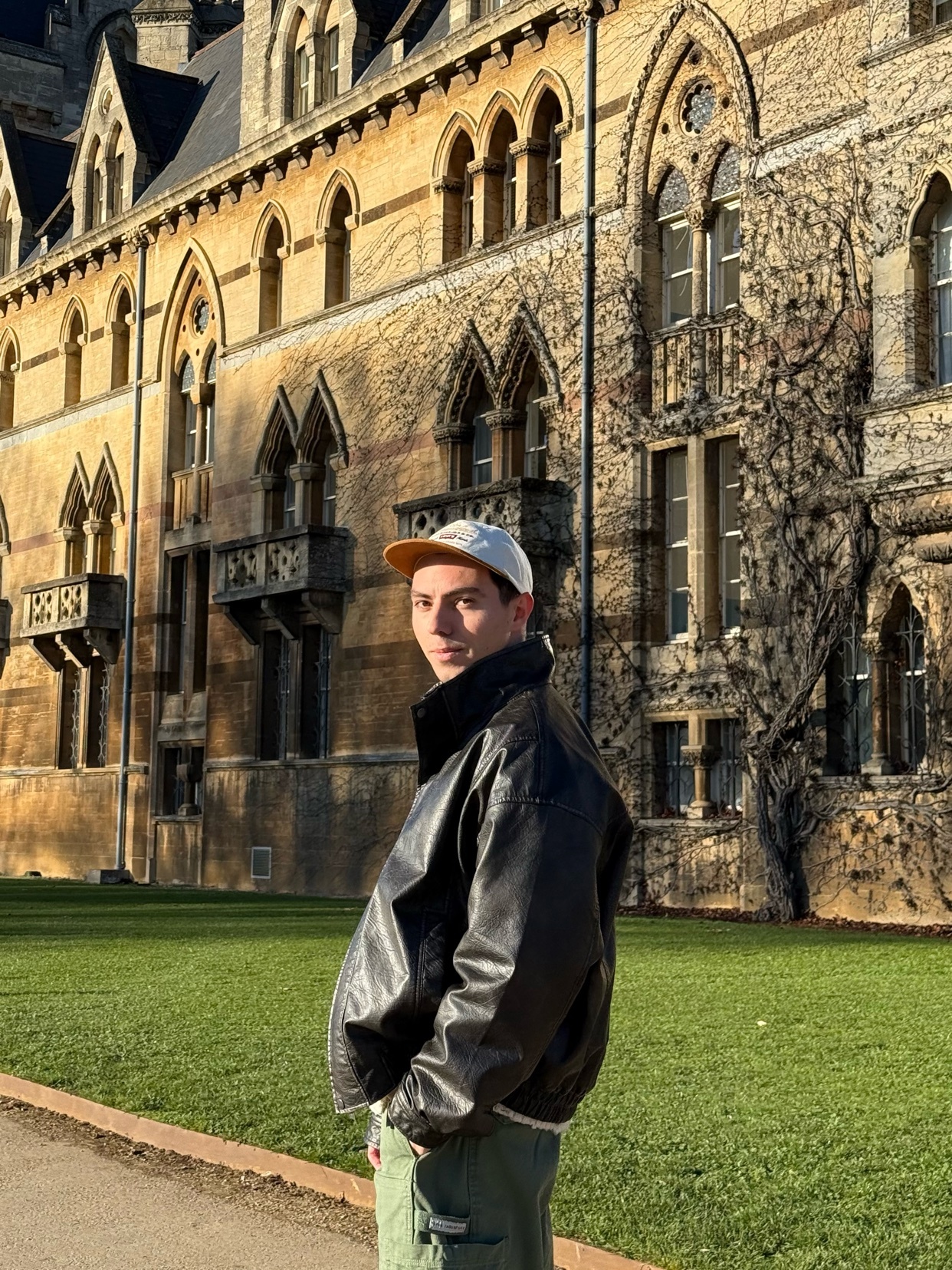 A man poses in front of a large, historic building. He is wearing a leather jacket and a cap.