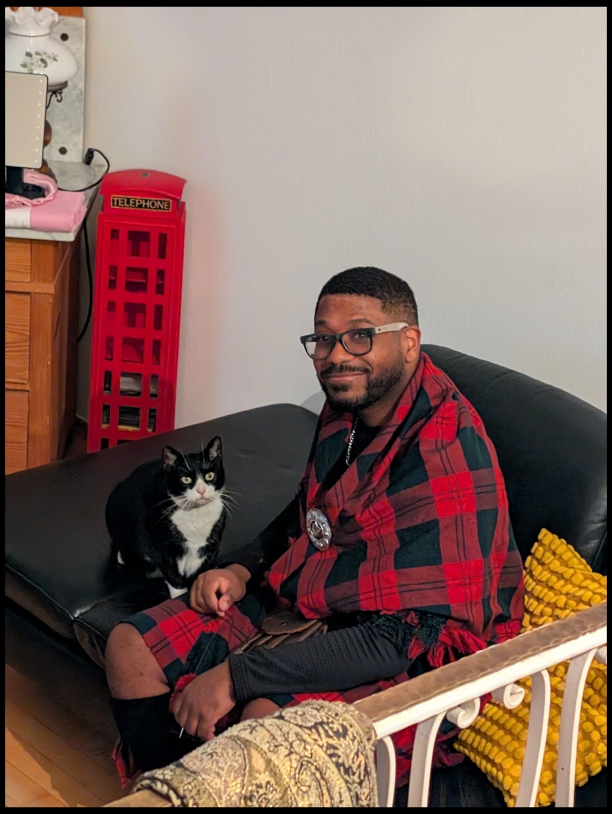 A man wearing a kilt and glasses sits on a couch next to a black and white cat. He is wrapped in a red and black plaid blanket.