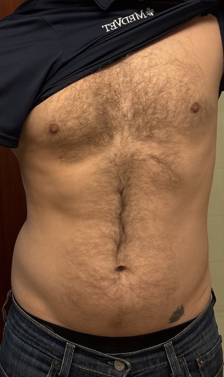 A man shows off his hairy chest and torso, wearing jeans and lifting his shirt.
