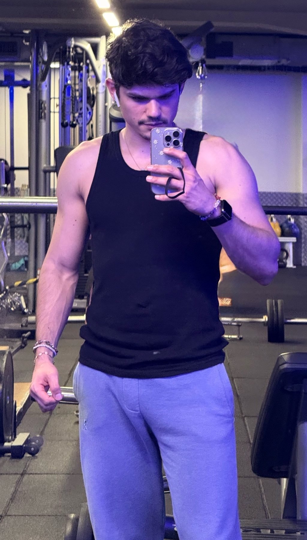 A man takes a mirror selfie in a gym, showcasing his physique. He's wearing a black tank top and gray sweatpants.