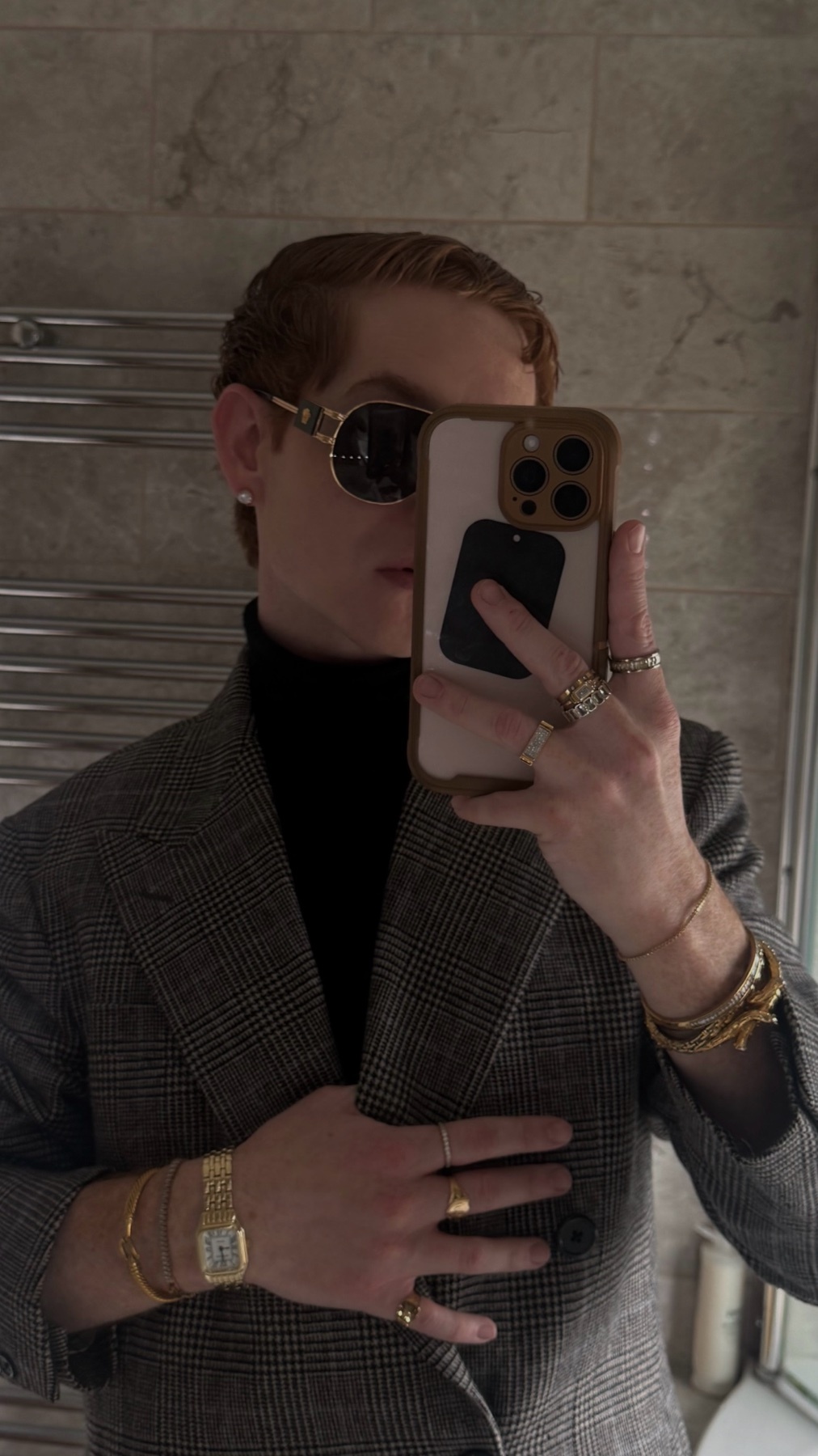 A man in sunglasses and a blazer takes a mirror selfie, accessorized with multiple rings and watches.