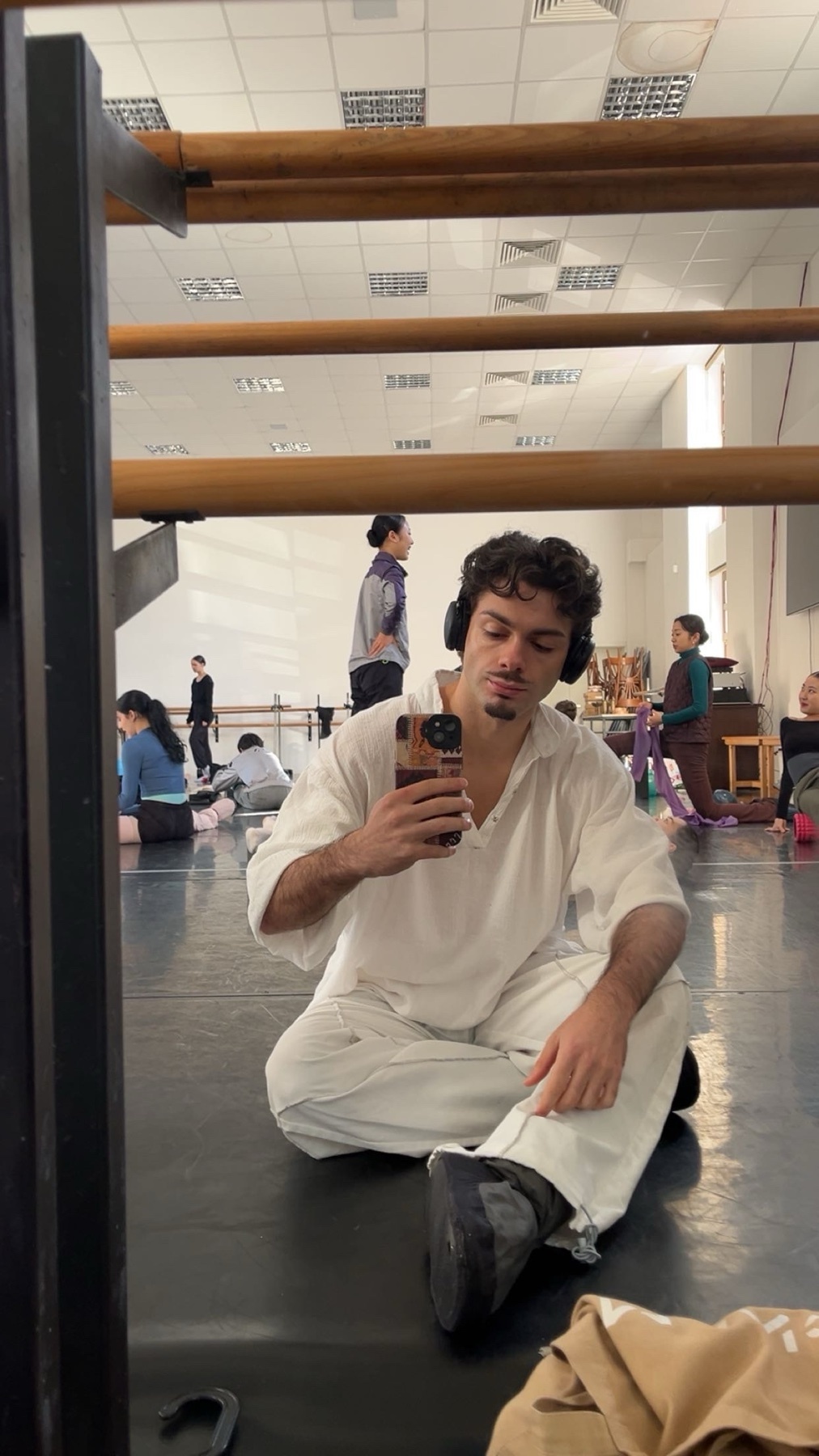 A man in white clothing sits on the floor of a dance studio, taking a selfie in a mirror while wearing headphones. Other dancers are visible in the background, stretching and practicing.