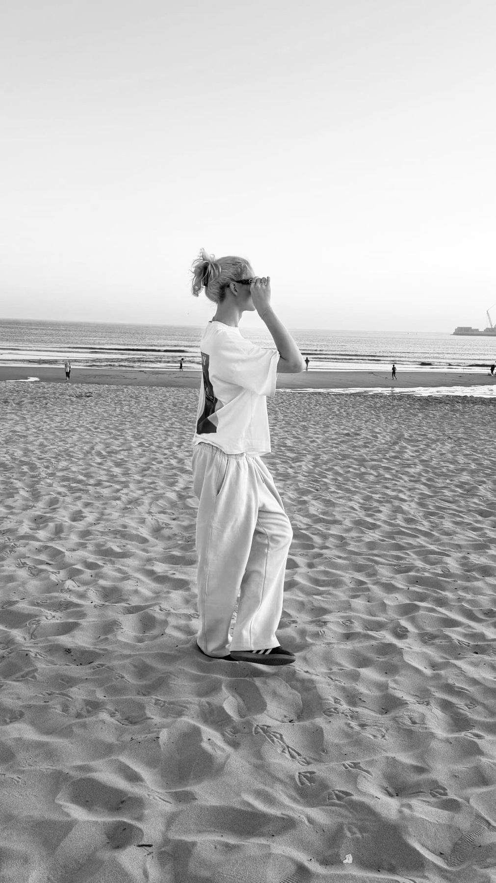 A black and white photo of a woman standing on a sandy beach in Porto, looking out at the ocean. She is wearing a t-shirt and pants, with her blonde hair pulled back in a ponytail.