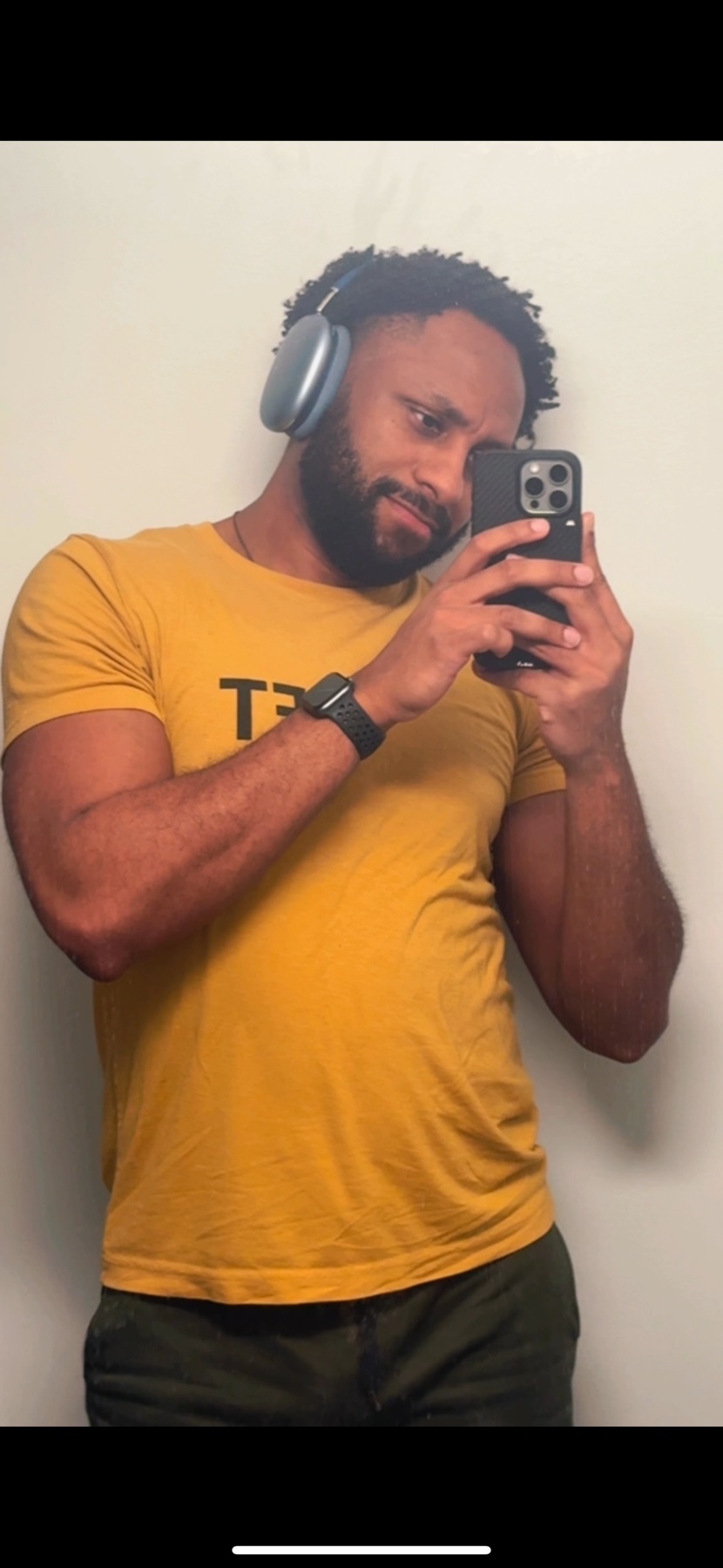 A man takes a selfie while wearing headphones and a yellow t-shirt. He has a beard and is looking at the camera.