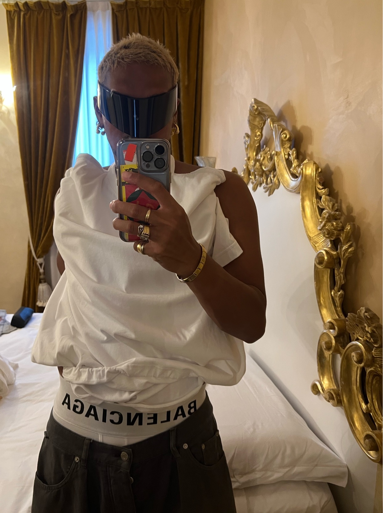 A woman takes a selfie in a room with a large ornate headboard. She is wearing a white t-shirt, dark pants, and large black sunglasses.